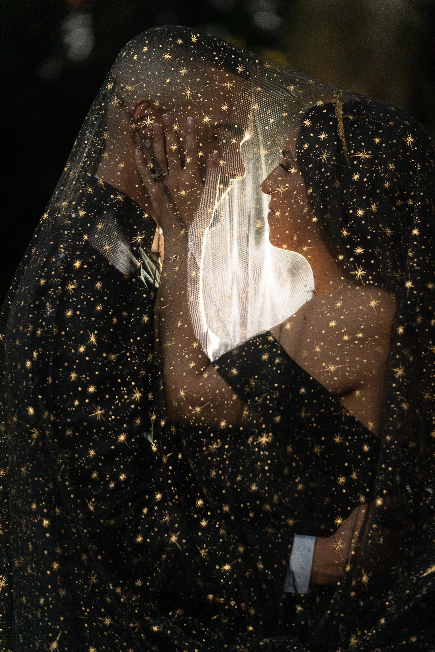 A couple, dressed in dark clothing, gently touching foreheads and gazing into each other's eyes under a transparent veil with gold star patterns, illuminated with soft light.