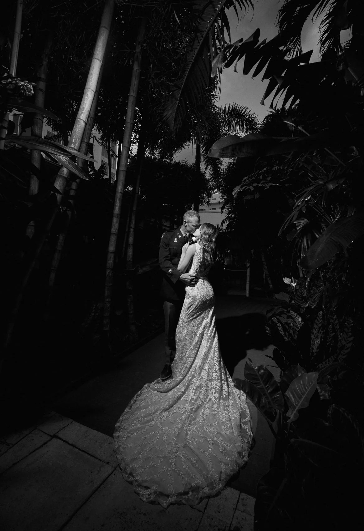 A black and white photo of a couple, with the man in a military uniform and the woman in a long, intricate wedding gown, sharing an intimate moment in a lush, tropical garden at night.