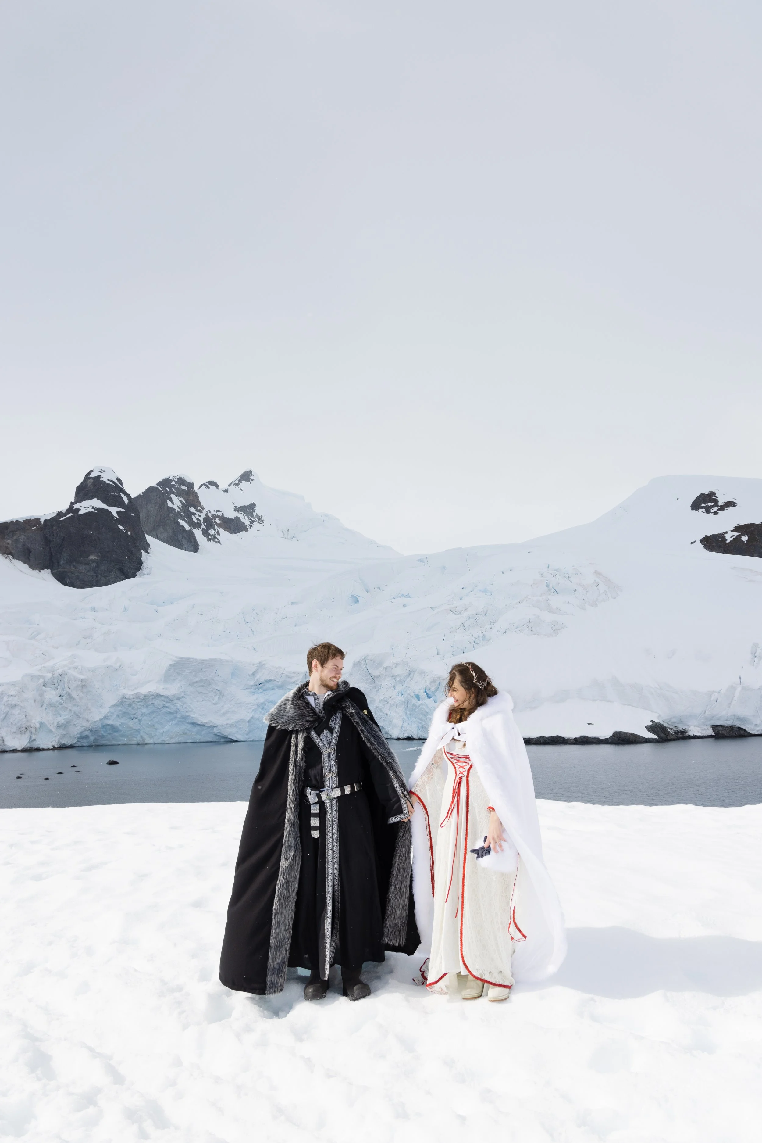 A couple dressed in traditional fantasy costumes standing on snow near a frozen lake with snow-covered mountains in the background.