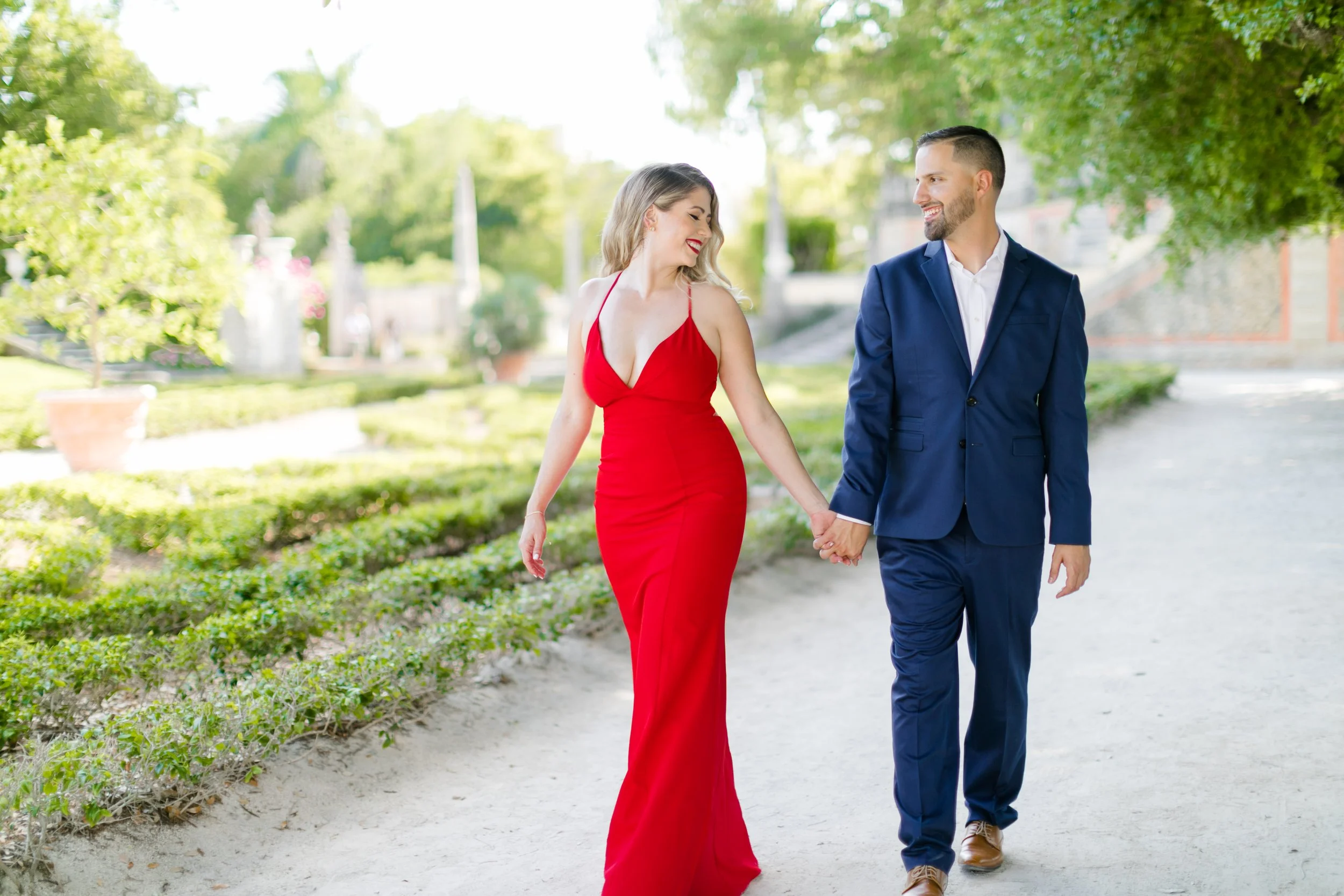 A couple walking hand in hand outdoors on a sunny day. The woman is wearing a red dress, and the man is wearing a navy suit.
