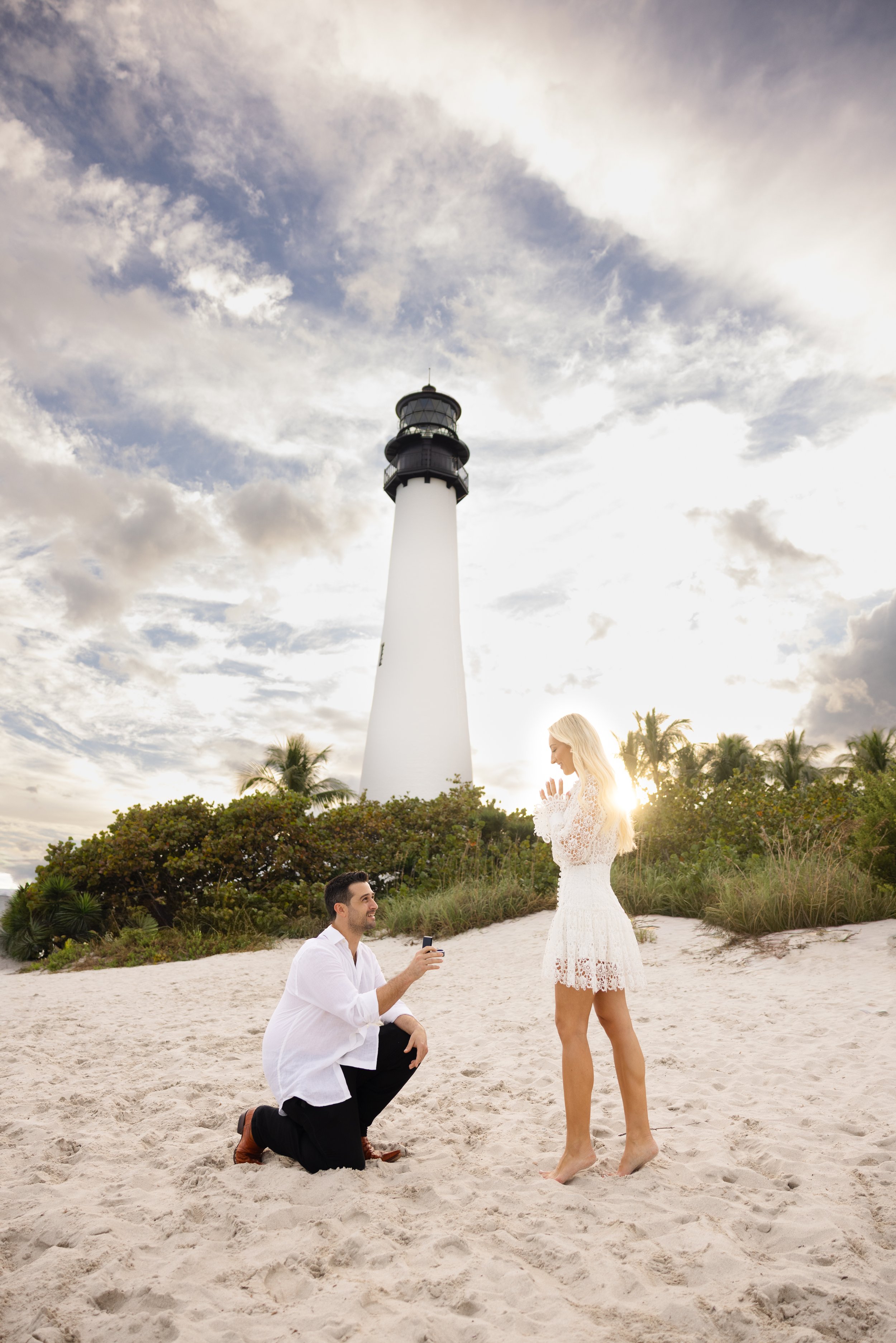 A man kneeling on the sandy beach proposing to a woman, with a lighthouse, trees, and cloudy sky in the background.