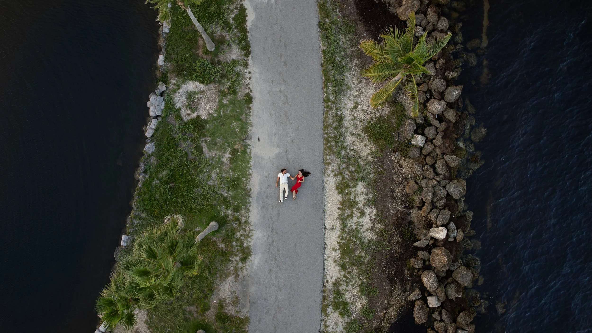 An aerial view of a couple lying on a gravel path between water bodies, with palm trees on either side.