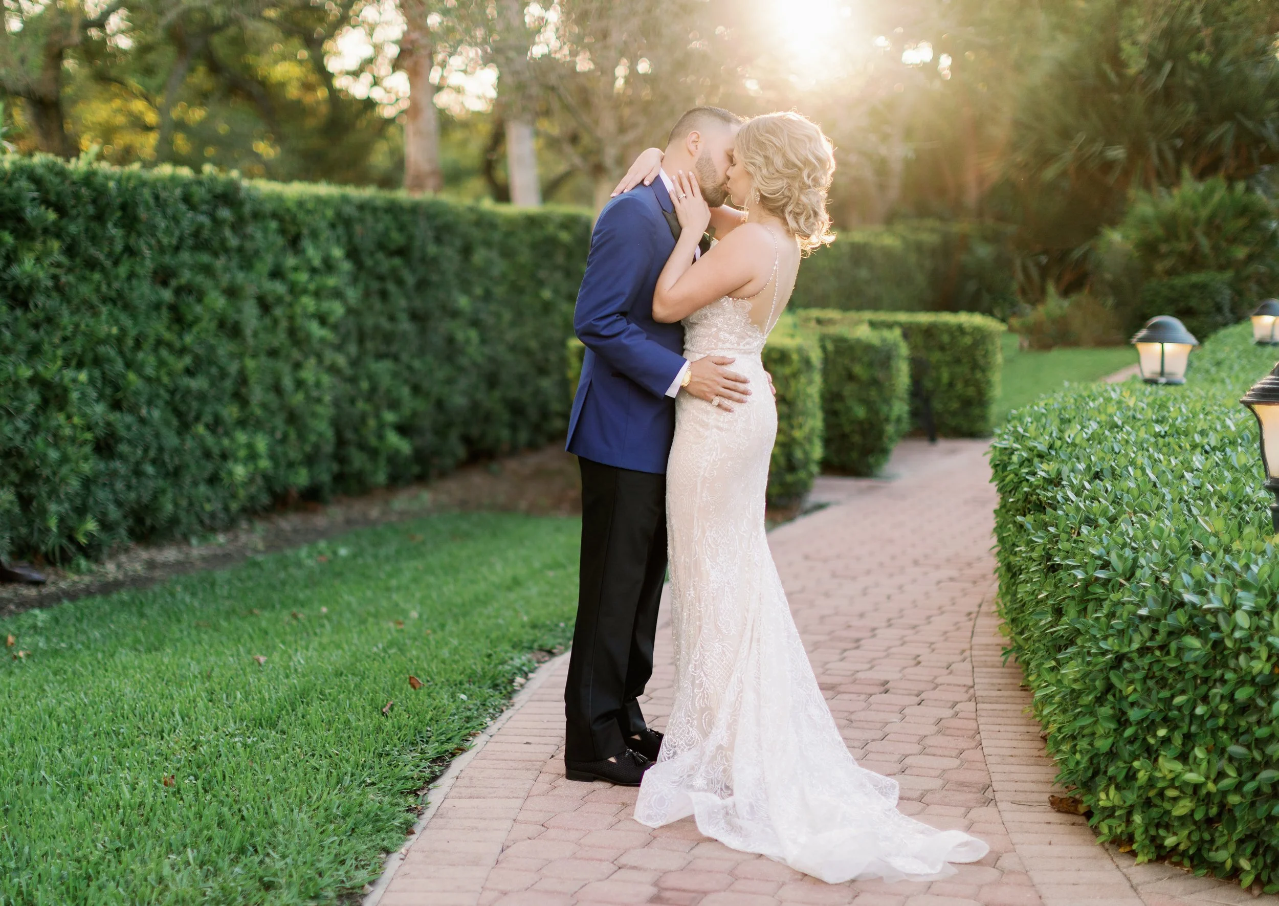 A bride and groom sharing a kiss outdoors on a brick pathway, surrounded by green bushes and trees, with sunlight shining through the trees in the background.
