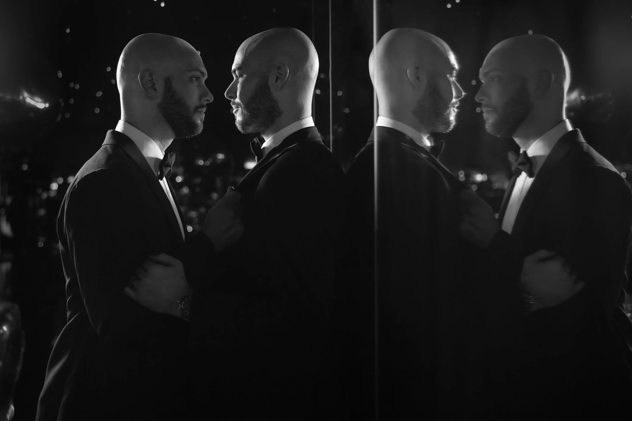 Two men dressed in tuxedos facing each other, mirrored in a glass, with one touching the other's chest, at night with city lights in the background.