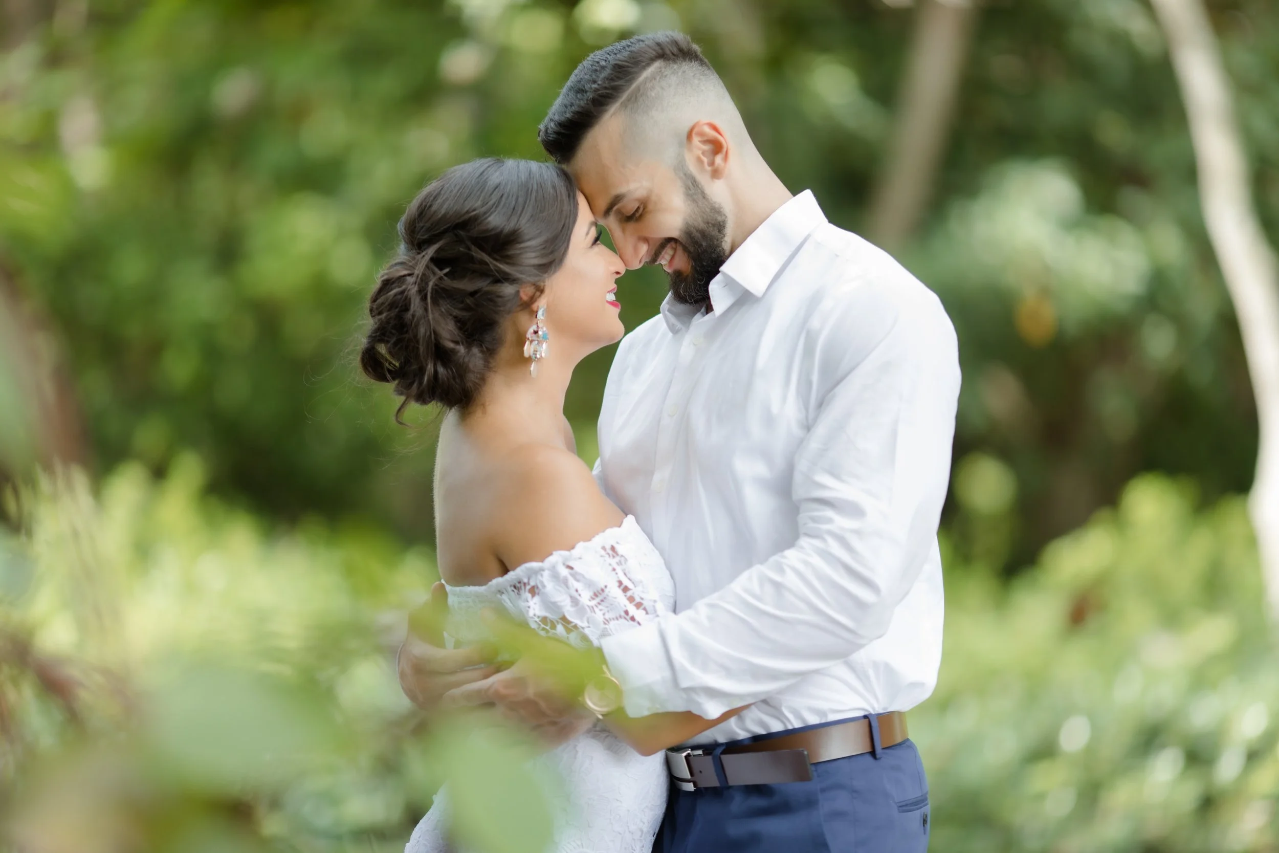 A couple embracing outdoors, their foreheads touching, in a lush green setting.