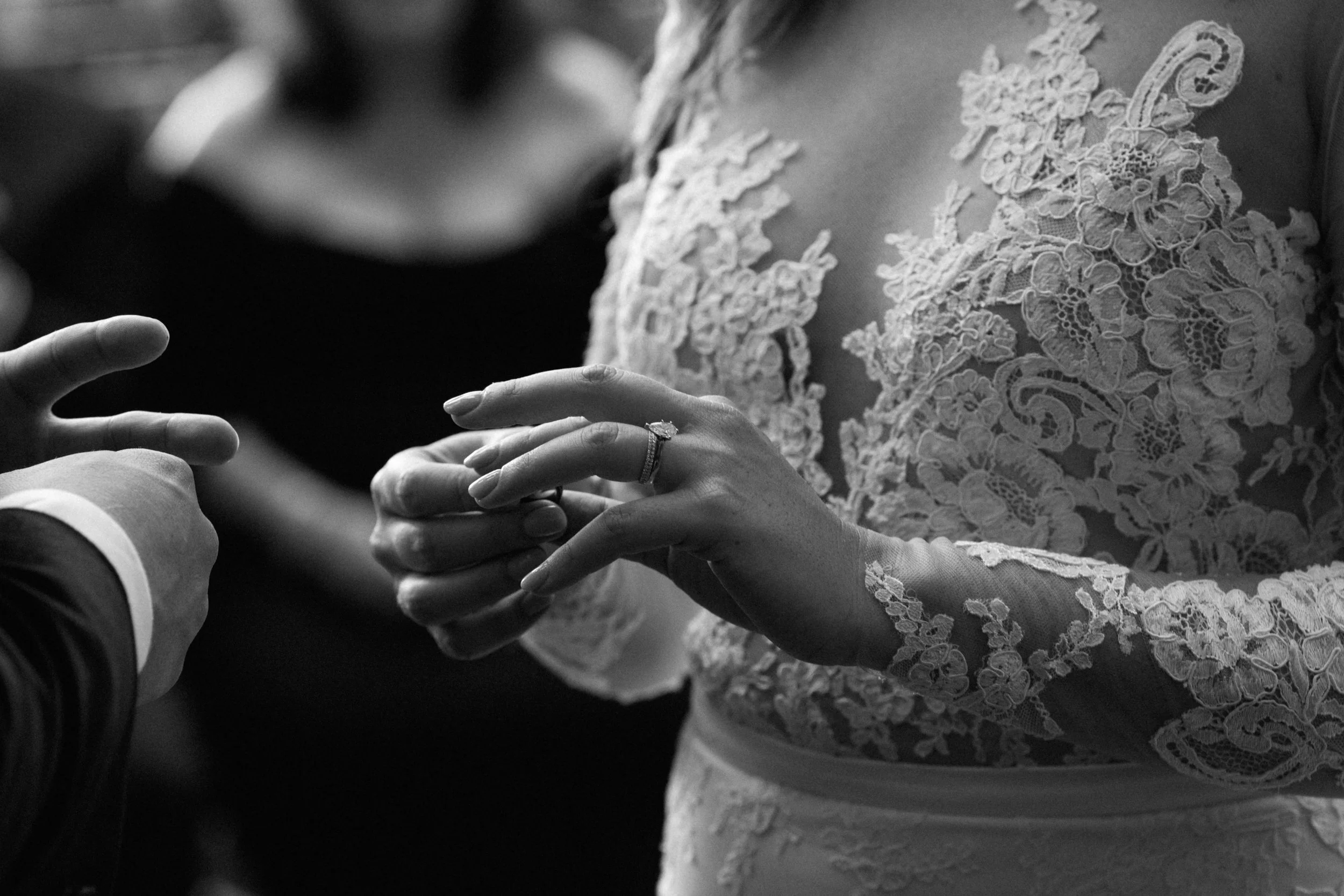 A bride wearing a lace wedding dress is holding hands with her groom during a wedding ceremony, with a focus on their hands and wedding ring.
