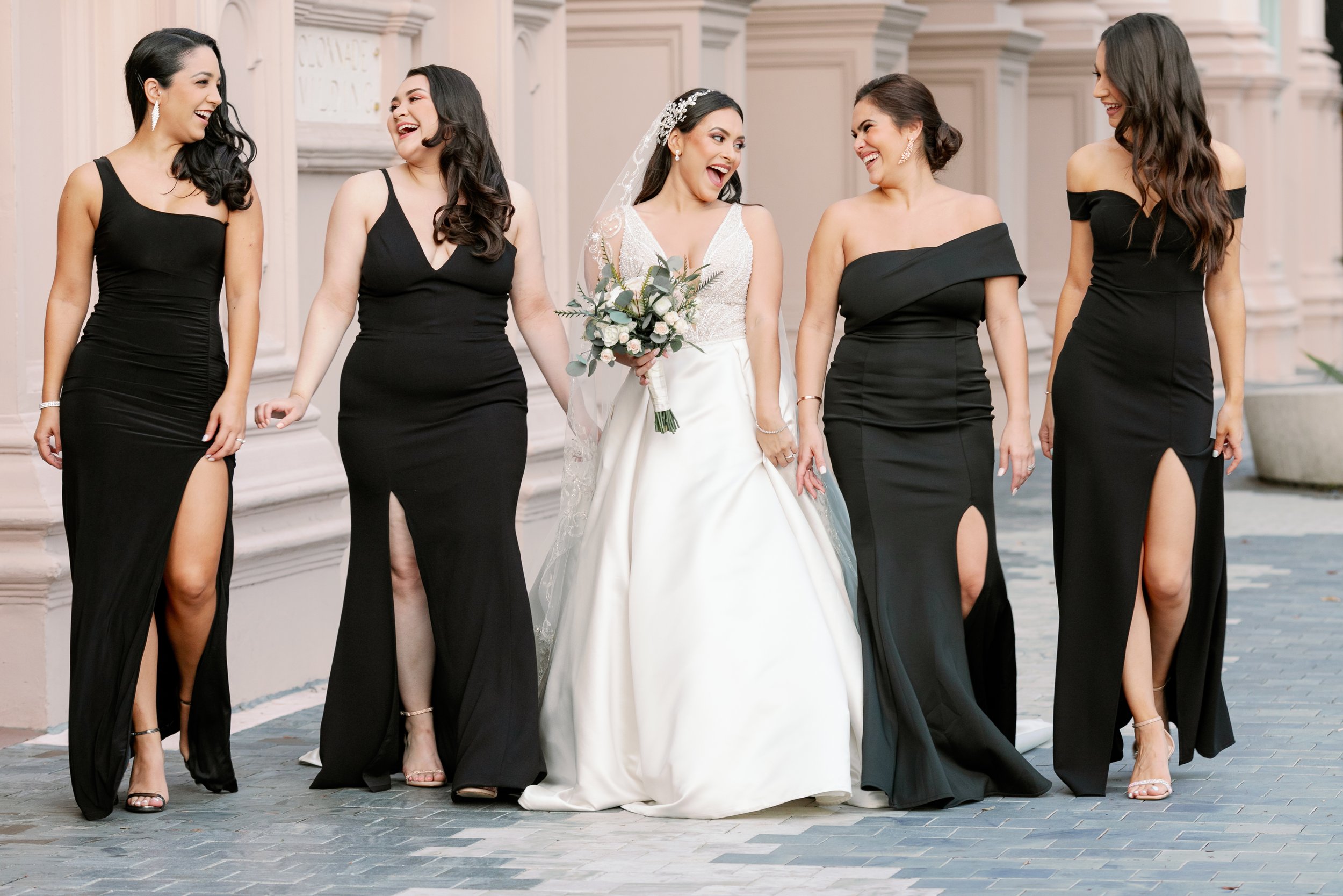 A bride in a white wedding dress with a bouquet walks with five bridesmaids in black dresses, all smiling and laughing outside a building.
