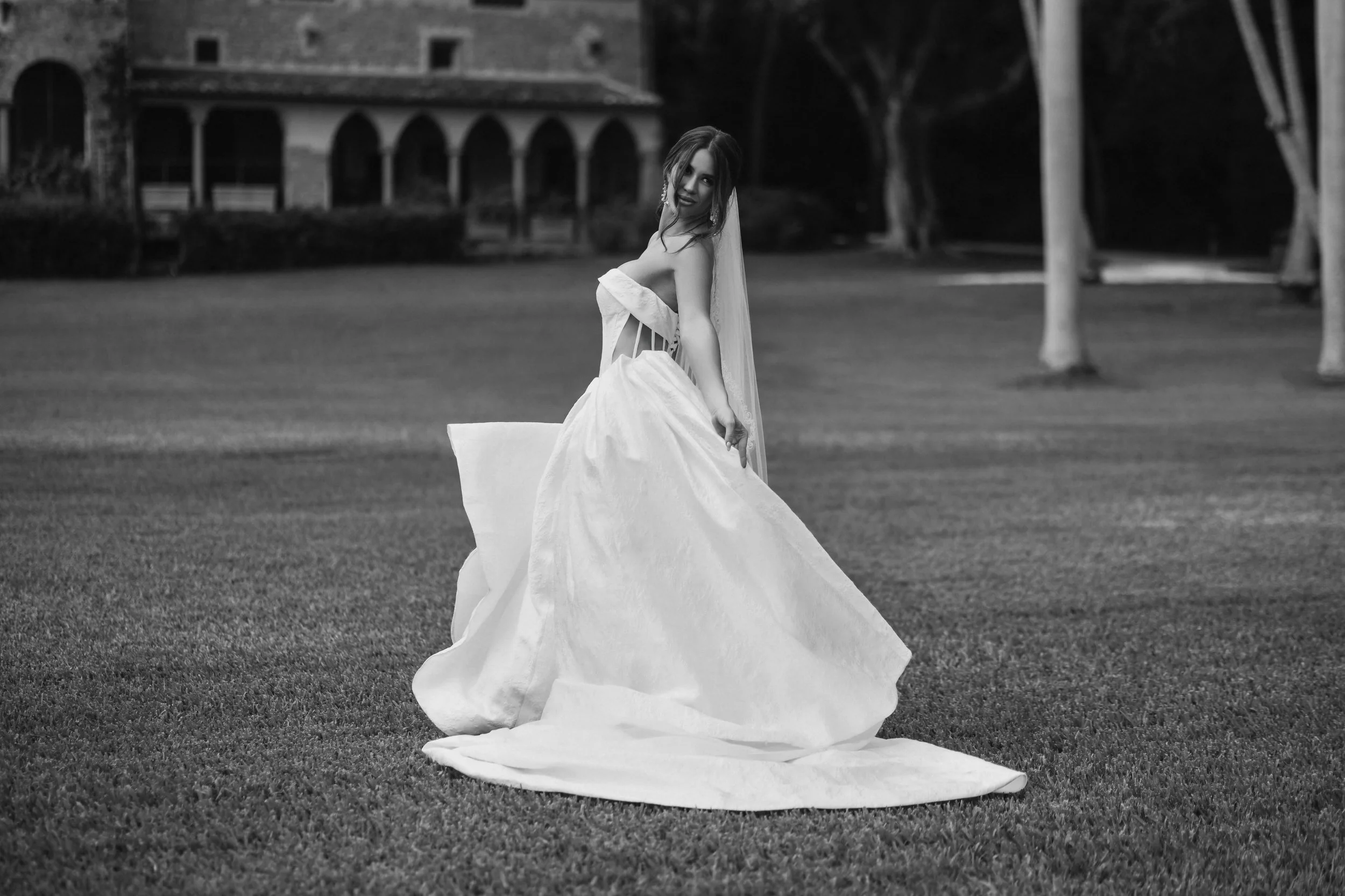 A woman in a wedding dress standing on a grass lawn, holding the skirt elegantly, with an old building and trees in the background.