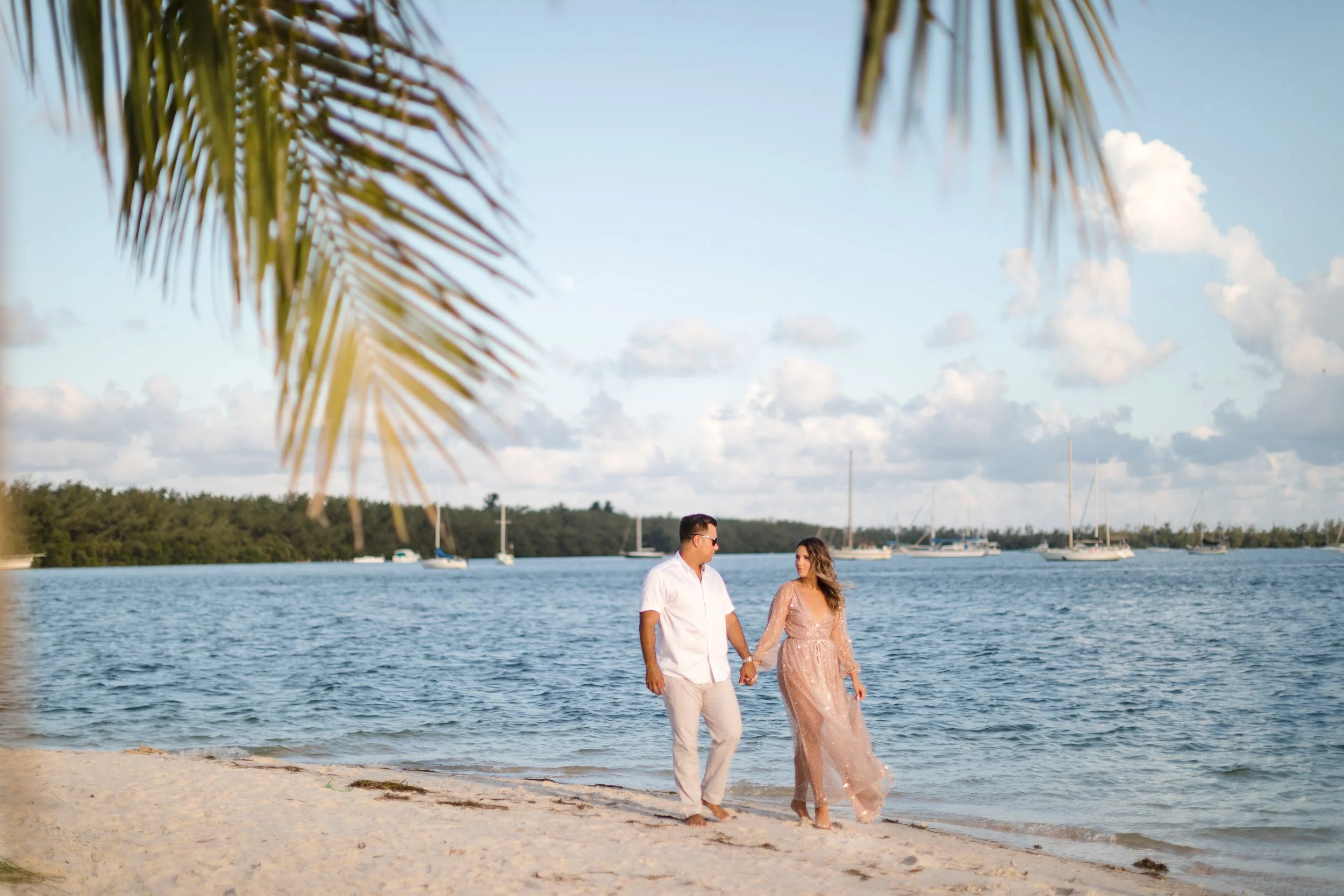 A couple holding hands and walking on a sandy beach near the water, with sailboats in the distance under a partly cloudy sky, framed by palm tree leaves.