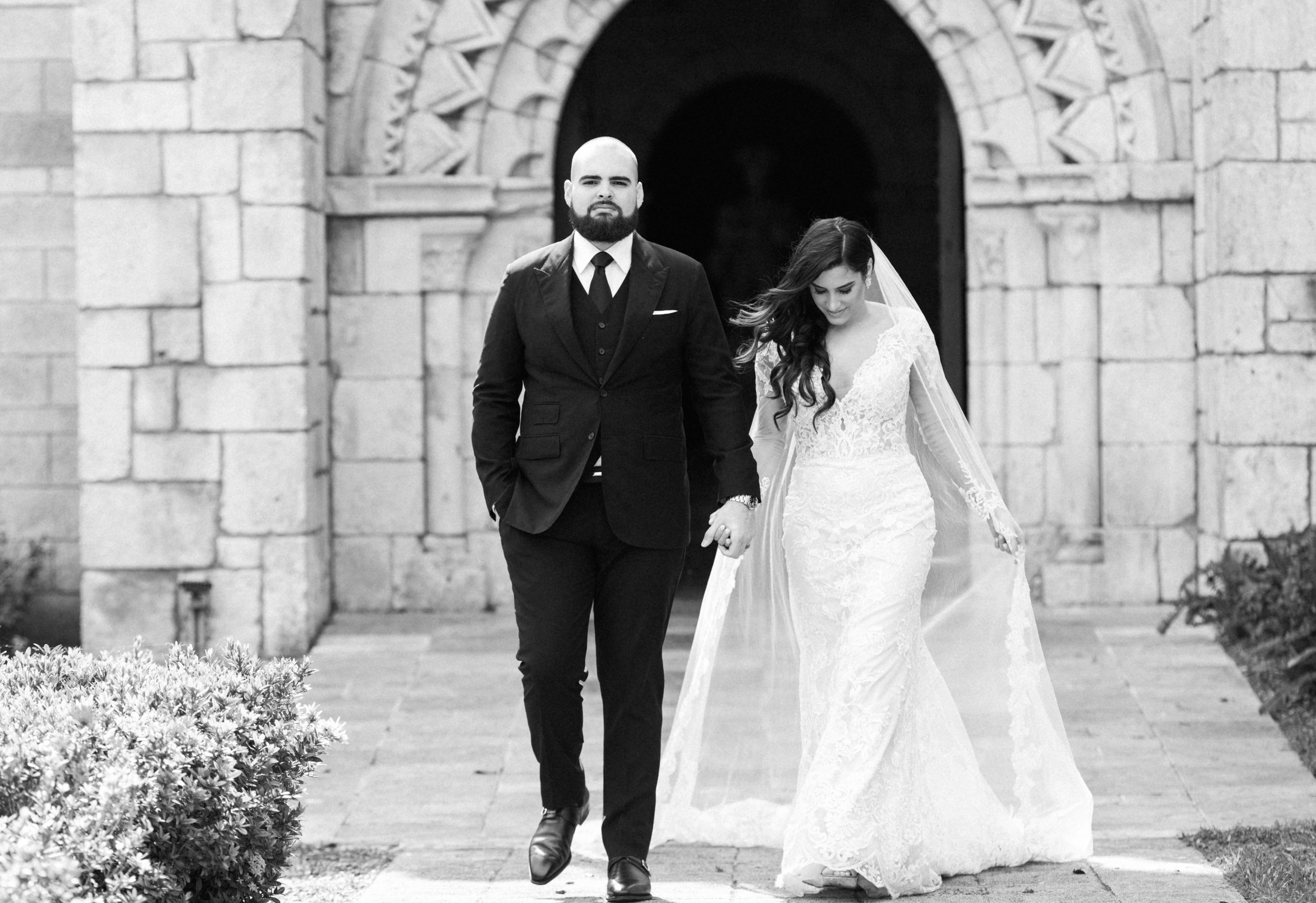 A bride and groom walking hand in hand outside a stone building, dressed in wedding attire, with the groom in a suit and the bride in a lace wedding gown, in black and white photo.