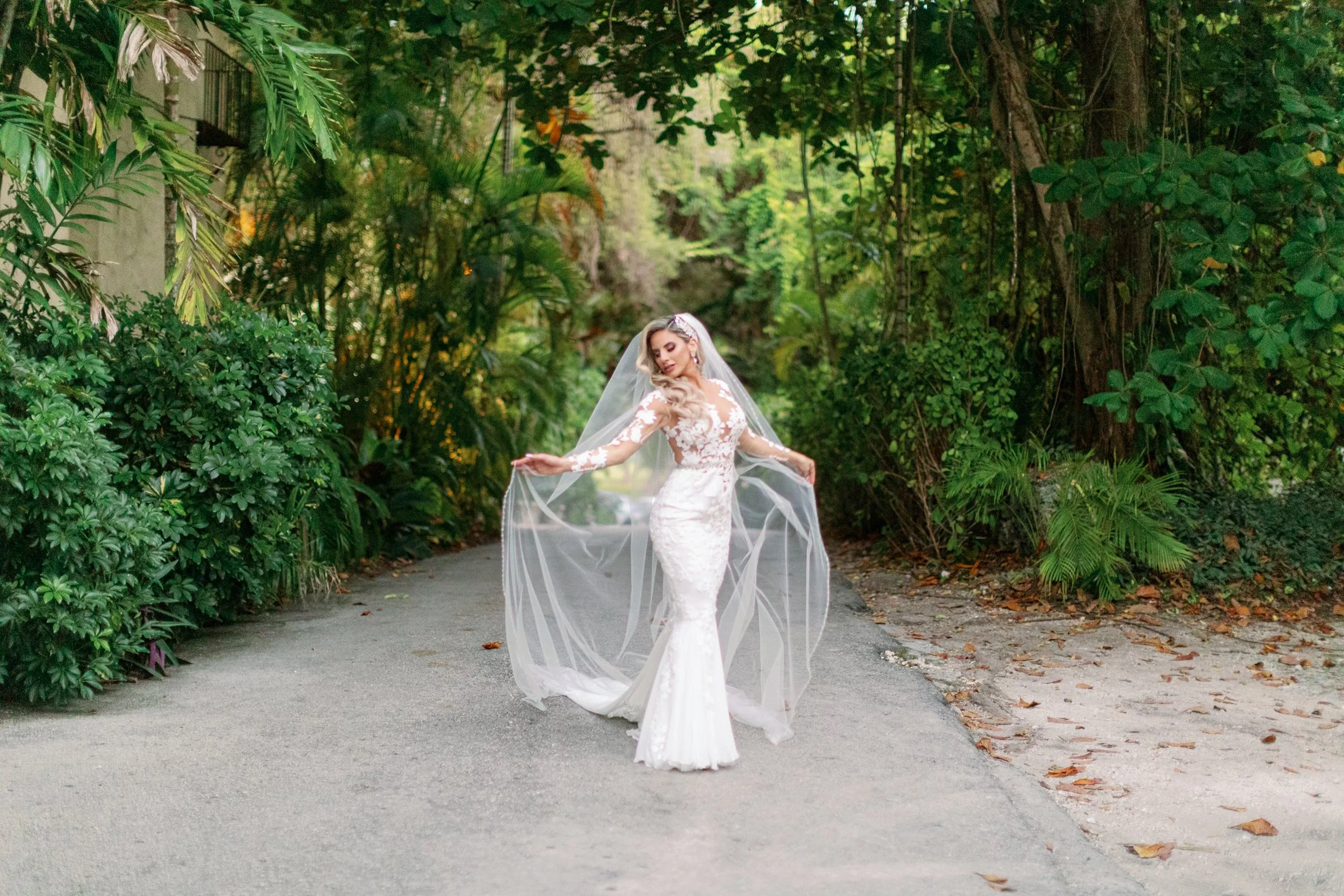 A bride in a white wedding gown with lace details poses on a wooded pathway, holding the edges of her veil in a lush green outdoor setting.