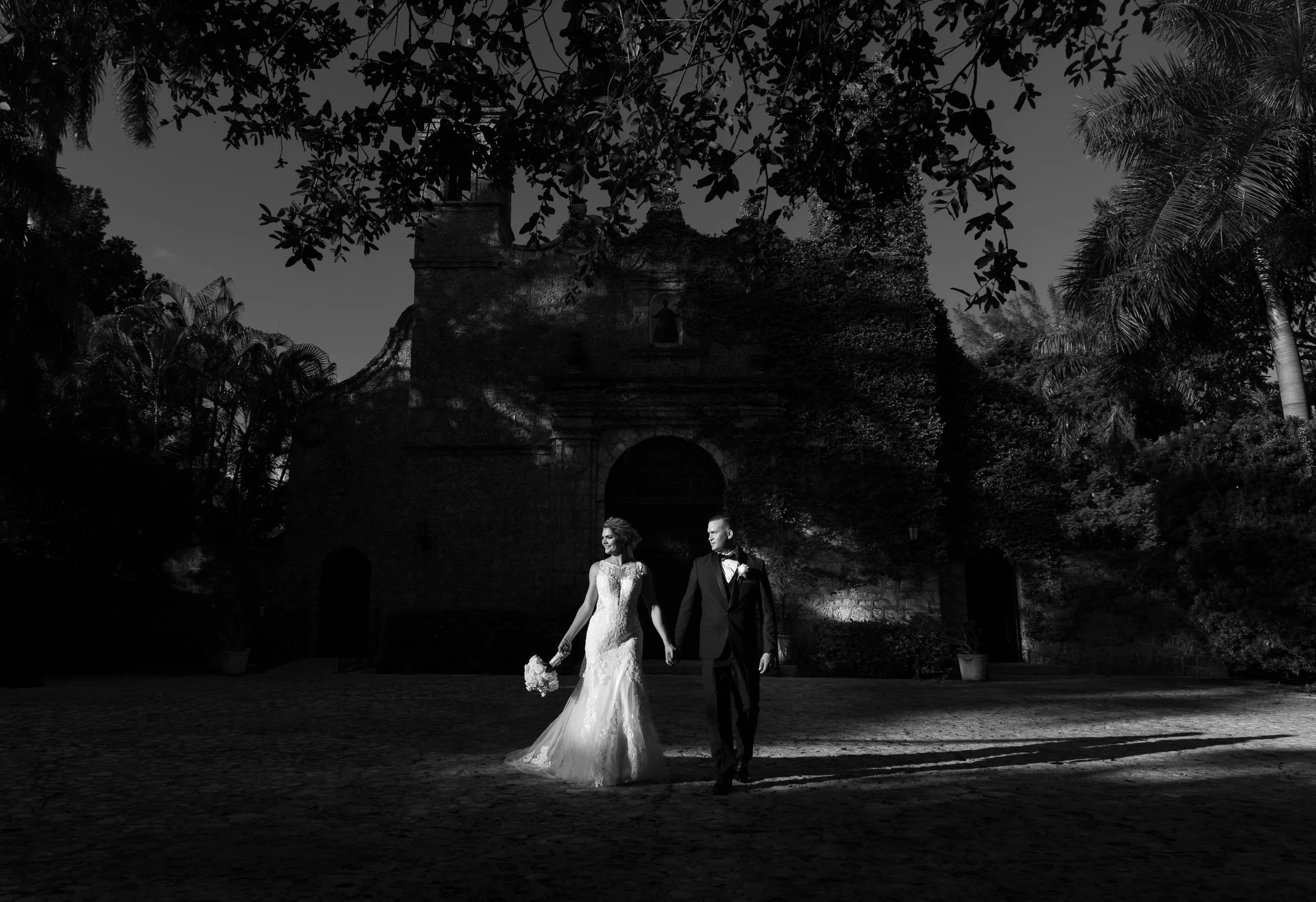 Black and white photo of a bride and groom holding hands, walking outdoors in front of an old stone building with surrounding trees, during what appears to be sunset.