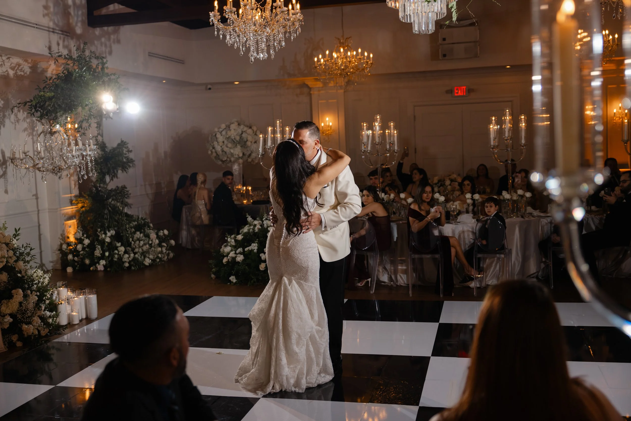 A bride and groom share their first dance at their wedding reception in a decorated banquet hall, with seated guests and large floral arrangements in the background.