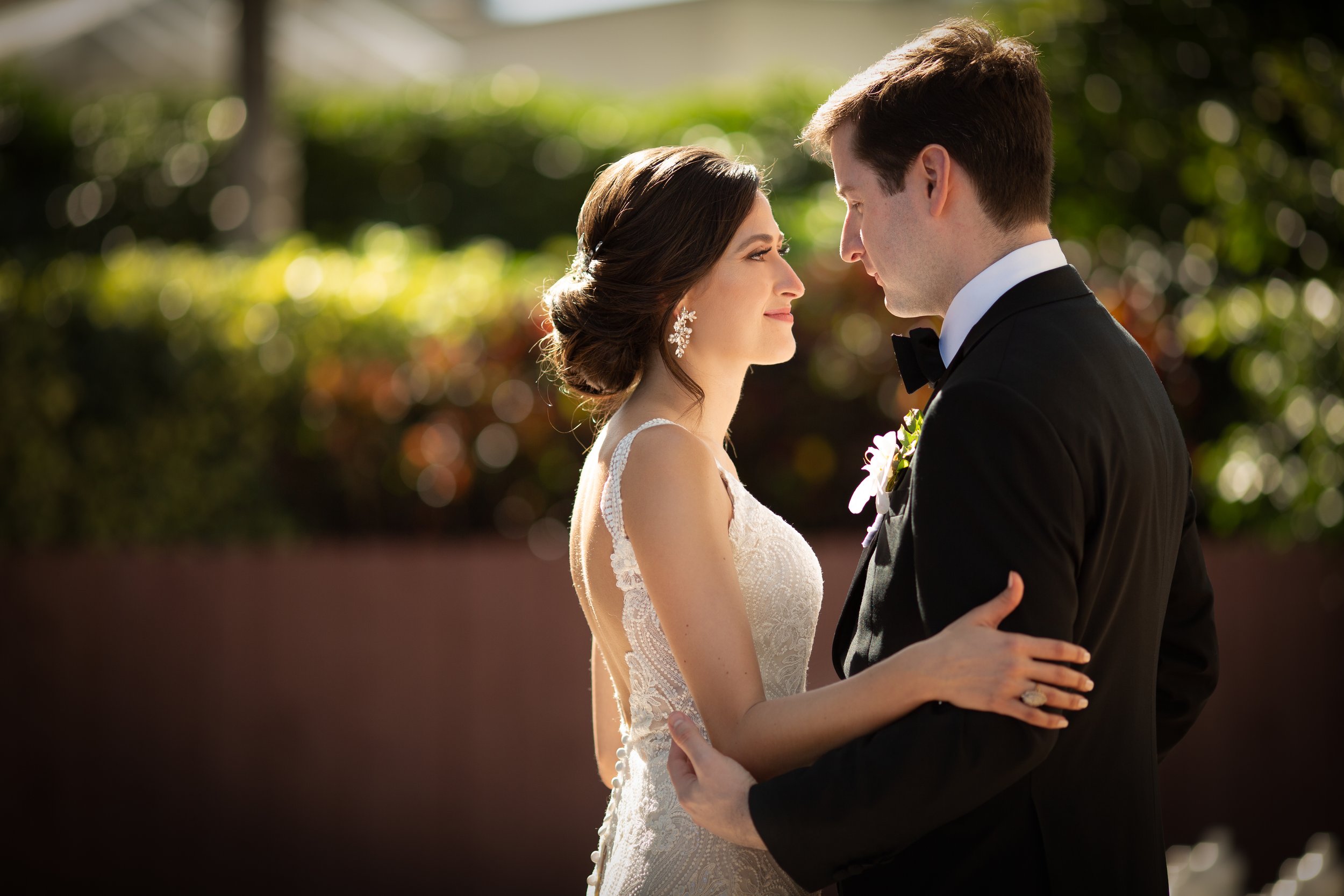 A bride and groom in wedding attire holding each other, standing close with foreheads touching in an outdoor setting with blurred greenery in the background.