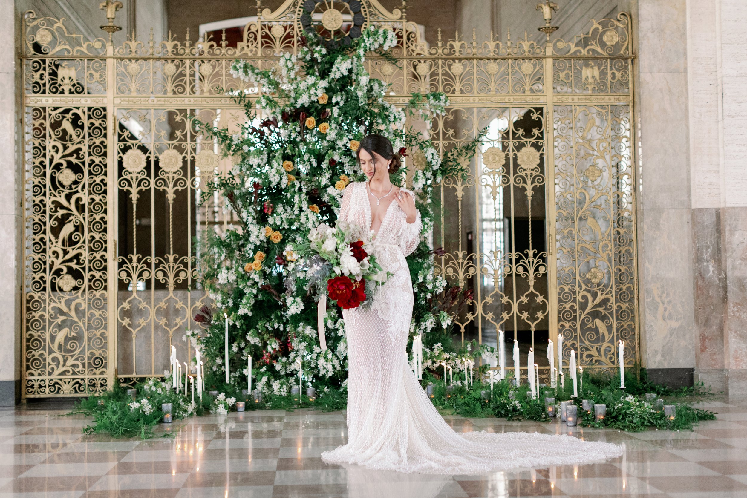 A bride in a white lace wedding gown stands in front of a large floral arrangement with white, red, and peach flowers, surrounded by candles on a green garland, in an elegant venue with ornate gold gate and marble floor.