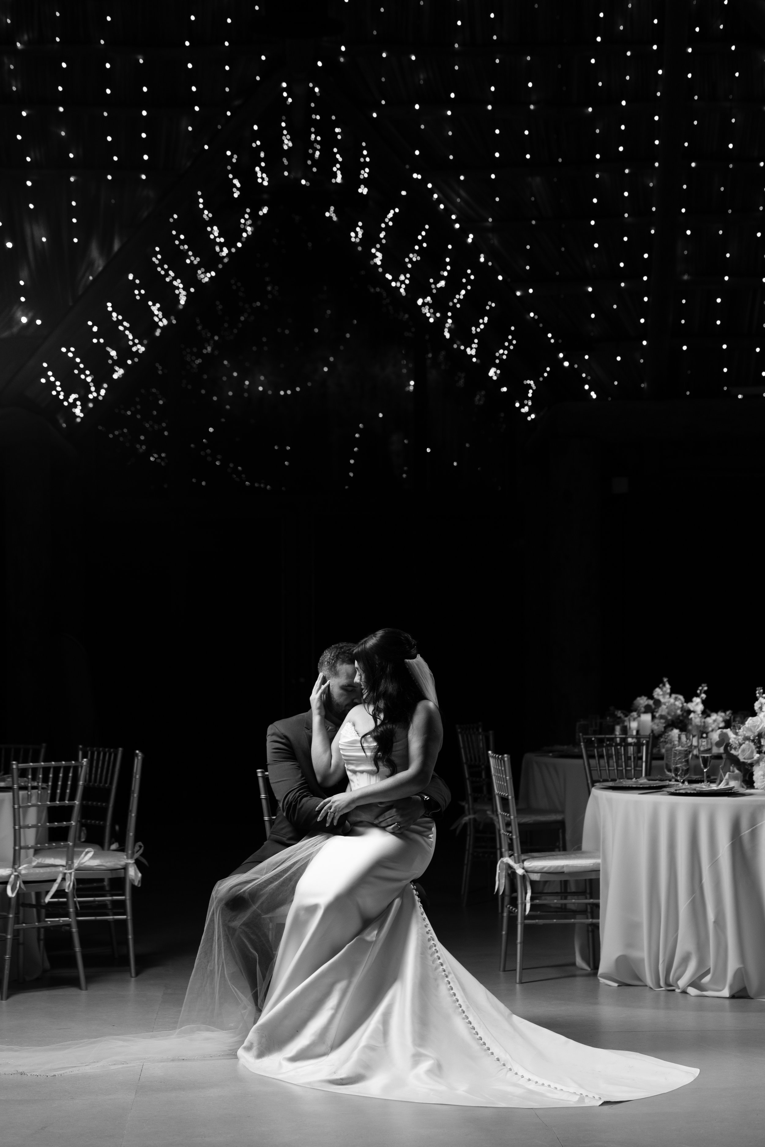 A black-and-white photo of a bride and groom sitting closely together in a wedding reception hall, under a canopy of string lights, with decorated tables and chairs in the background.