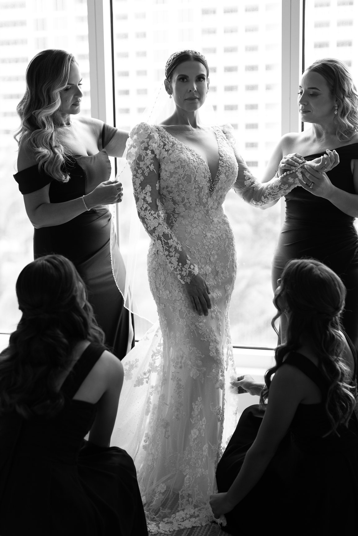 A bride in a lace wedding dress surrounded by her bridesmaids, with two of them kneeling and two standing, as they help her get ready in front of a large window in a cityscape.