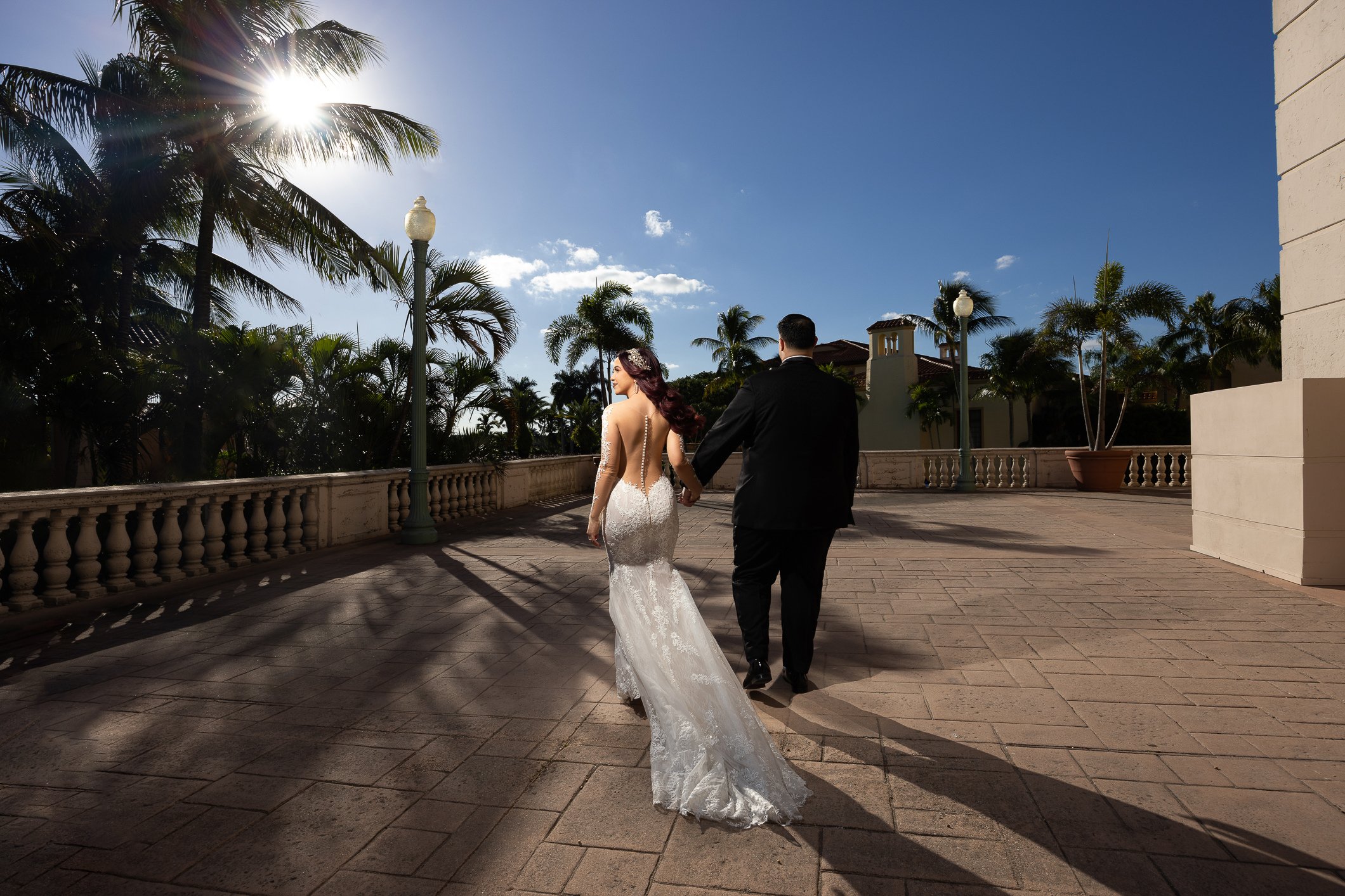 A bride and groom walking hand in hand outdoors on a sunny day with palm trees in the background.