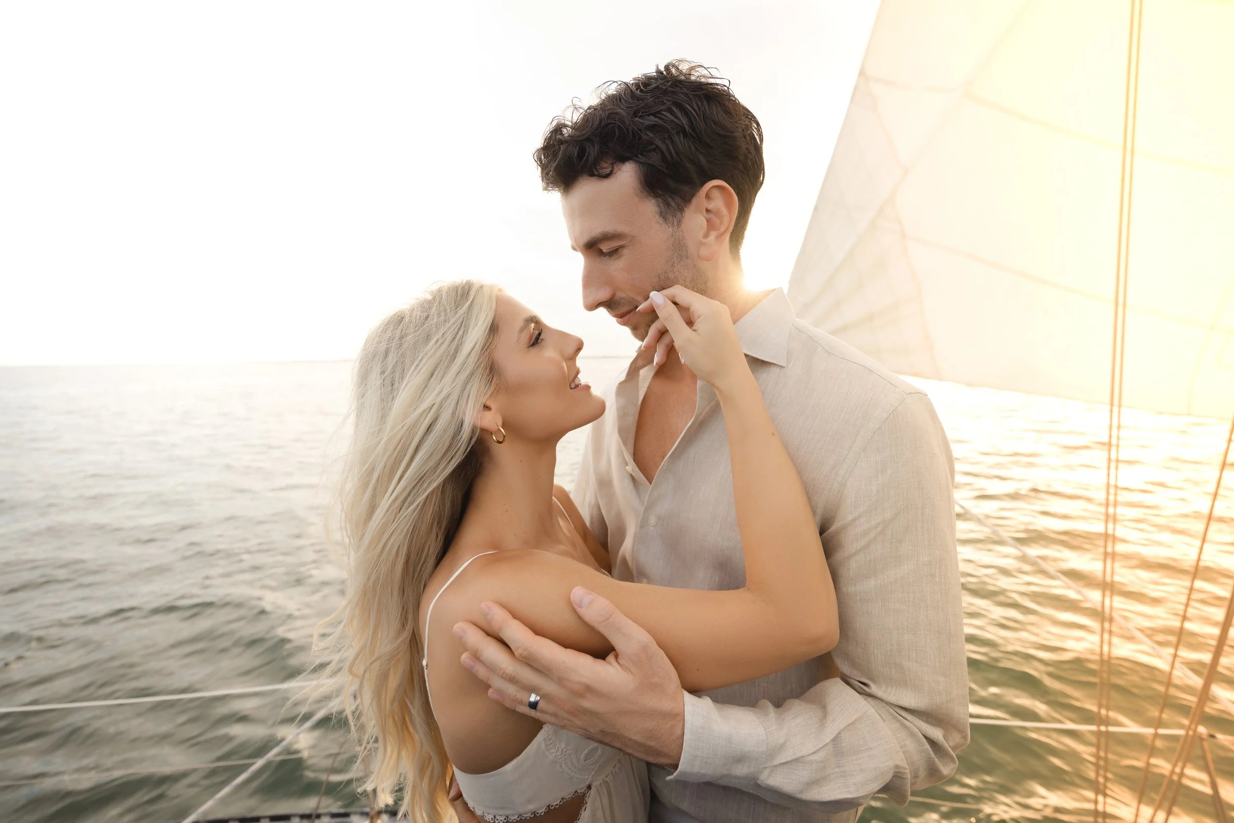A romantic couple with light skin embracing on a boat, looking into each other's eyes at sunset over the water.
