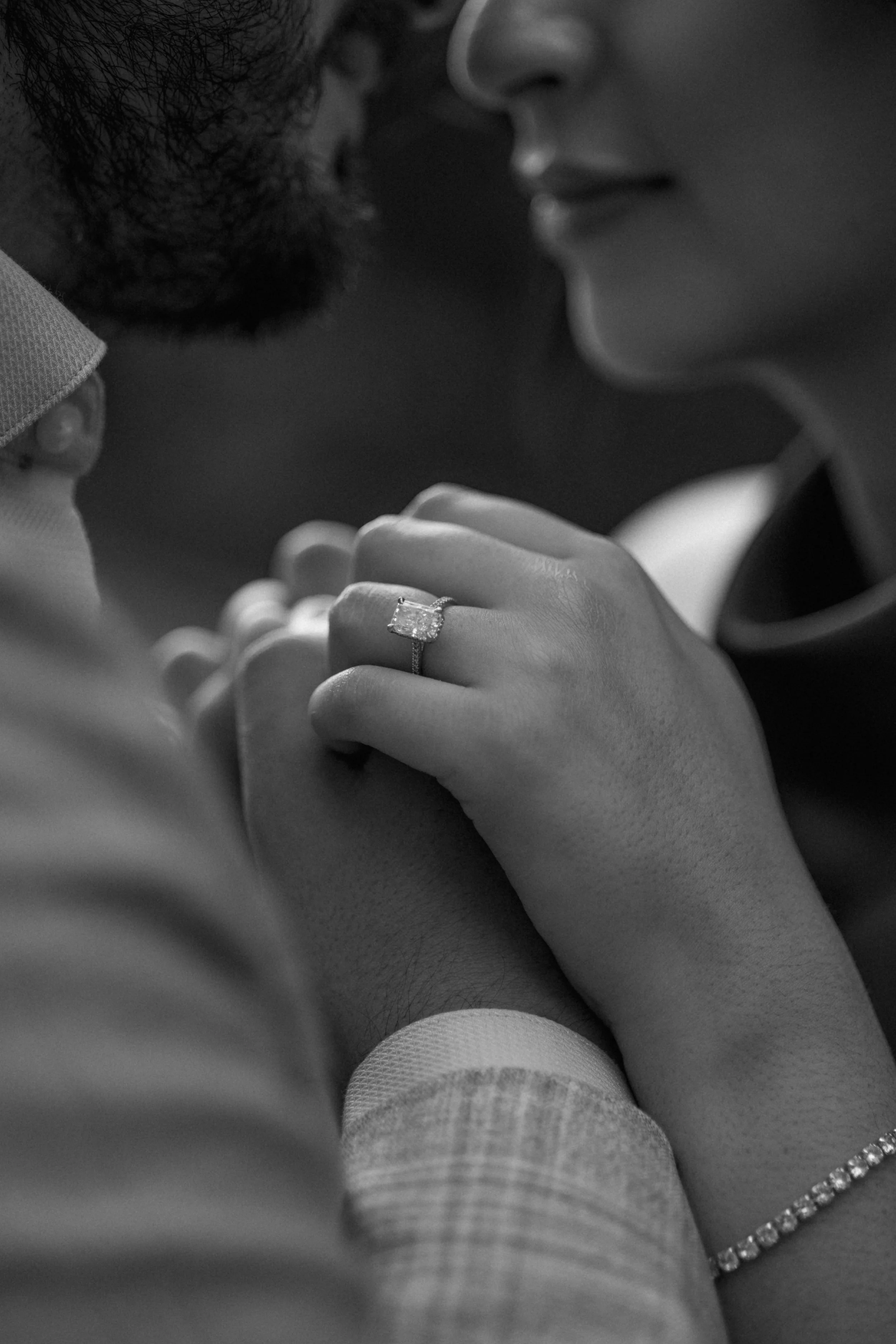 Close-up of a couple touching foreheads, with a focus on the woman's hand showing an engagement ring with a large square-shaped diamond. The scene is in black and white, capturing an intimate moment.