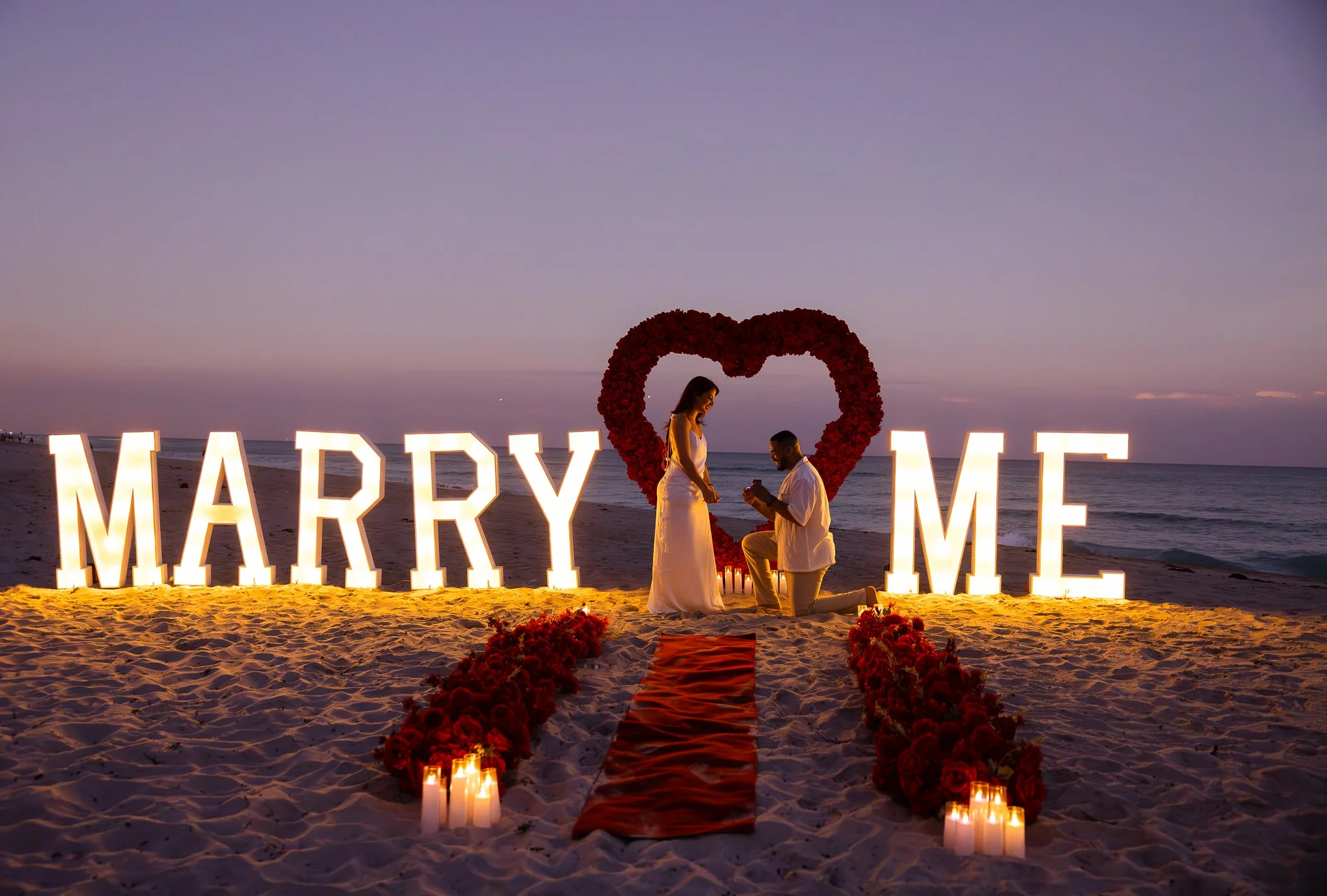 A couple on a beach during sunset, with large illuminated letters spelling 'MARRY ME' and a heart-shaped floral arrangement behind them, candles and flowers on the sand.
