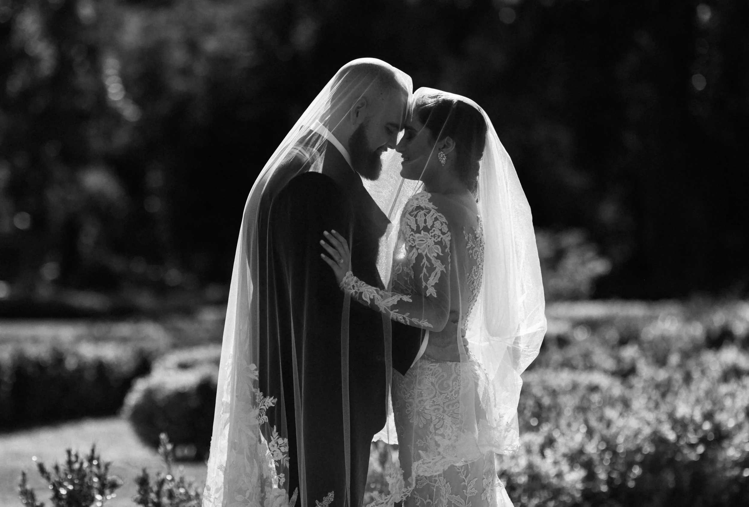 Black and white photograph of a bride and groom facing each other with foreheads touching, both wearing wedding veils, outdoors with blurred trees and foliage in the background.