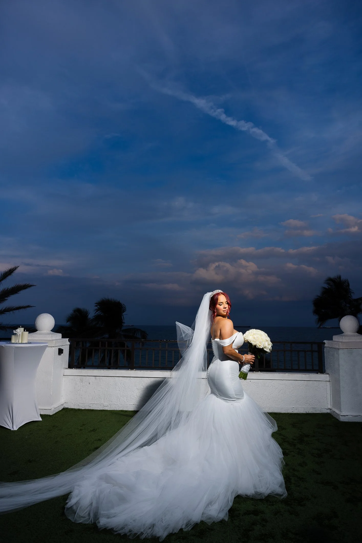 A bride in a white wedding gown with a long train and veil, holding a bouquet of white roses, standing outdoors on a terrace with a view of the ocean and sky.