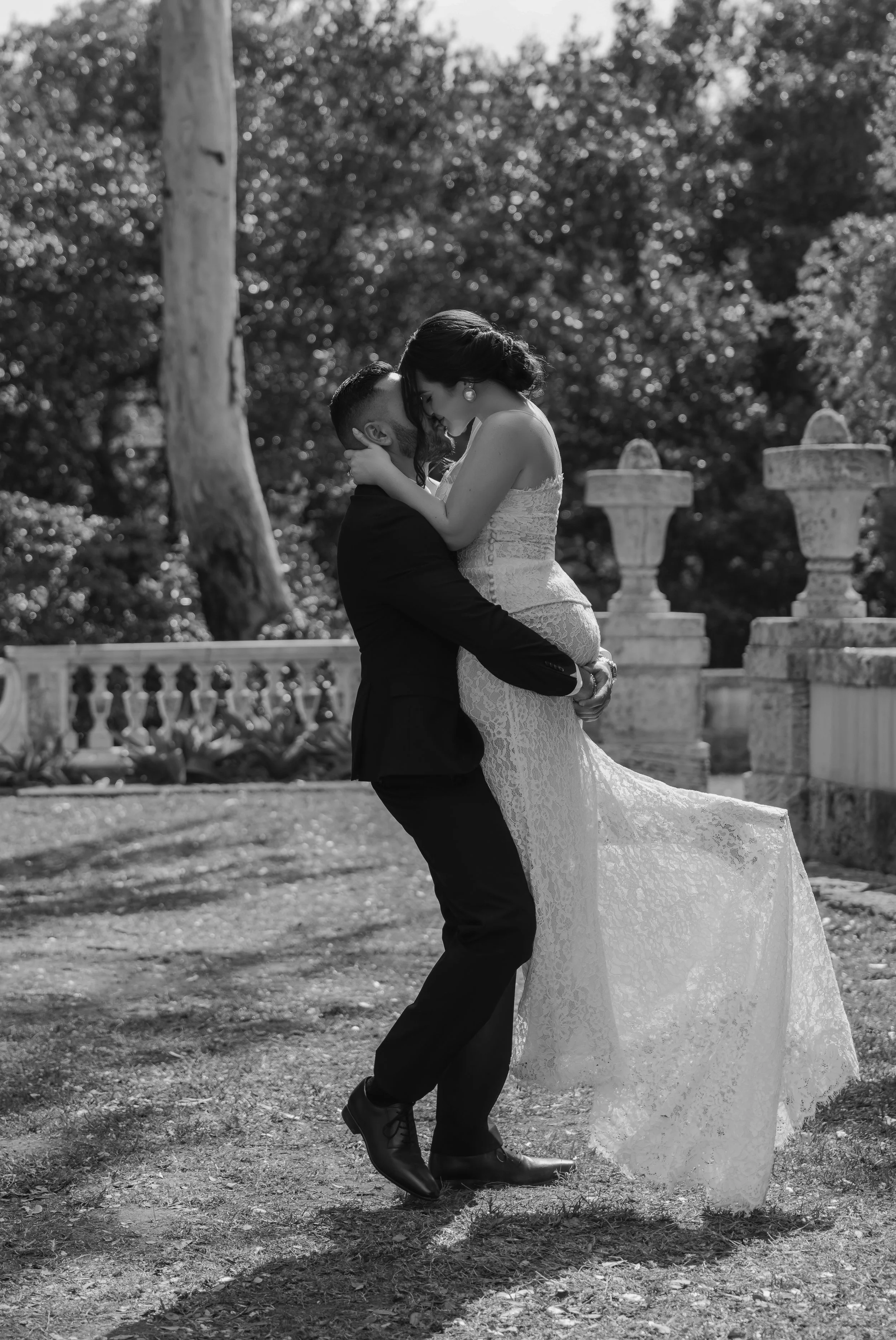 A couple in wedding attire embracing outdoors in black and white, with trees and stone urns in the background.