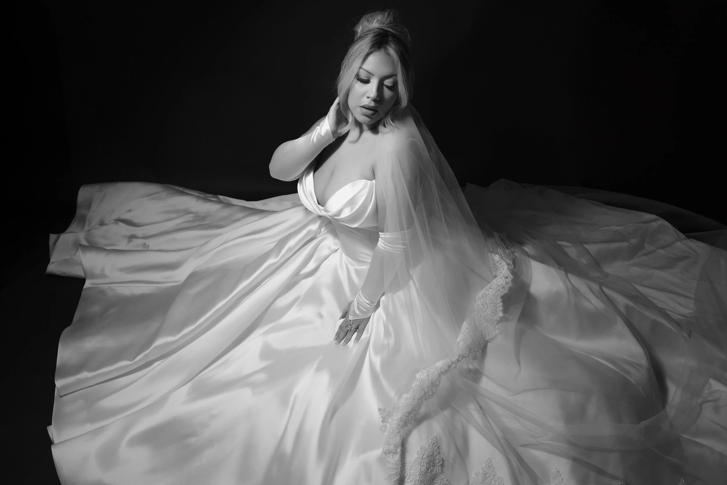 Black and white photo of a woman in a bridal gown sitting on the floor, with a dark background.