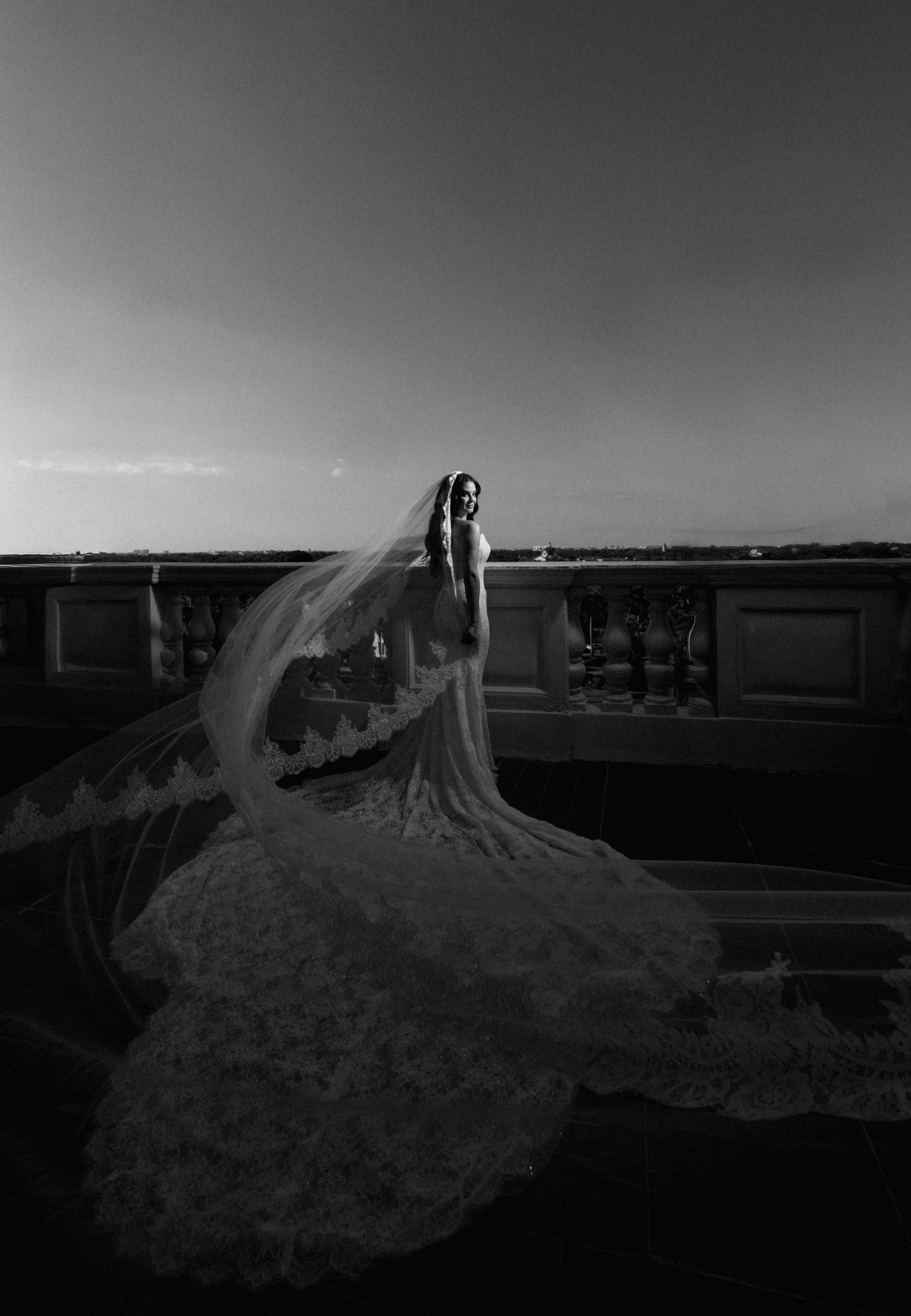 Black and white photo of a woman in a wedding dress with a long train and veil, standing on a balcony with a cityscape in the background.