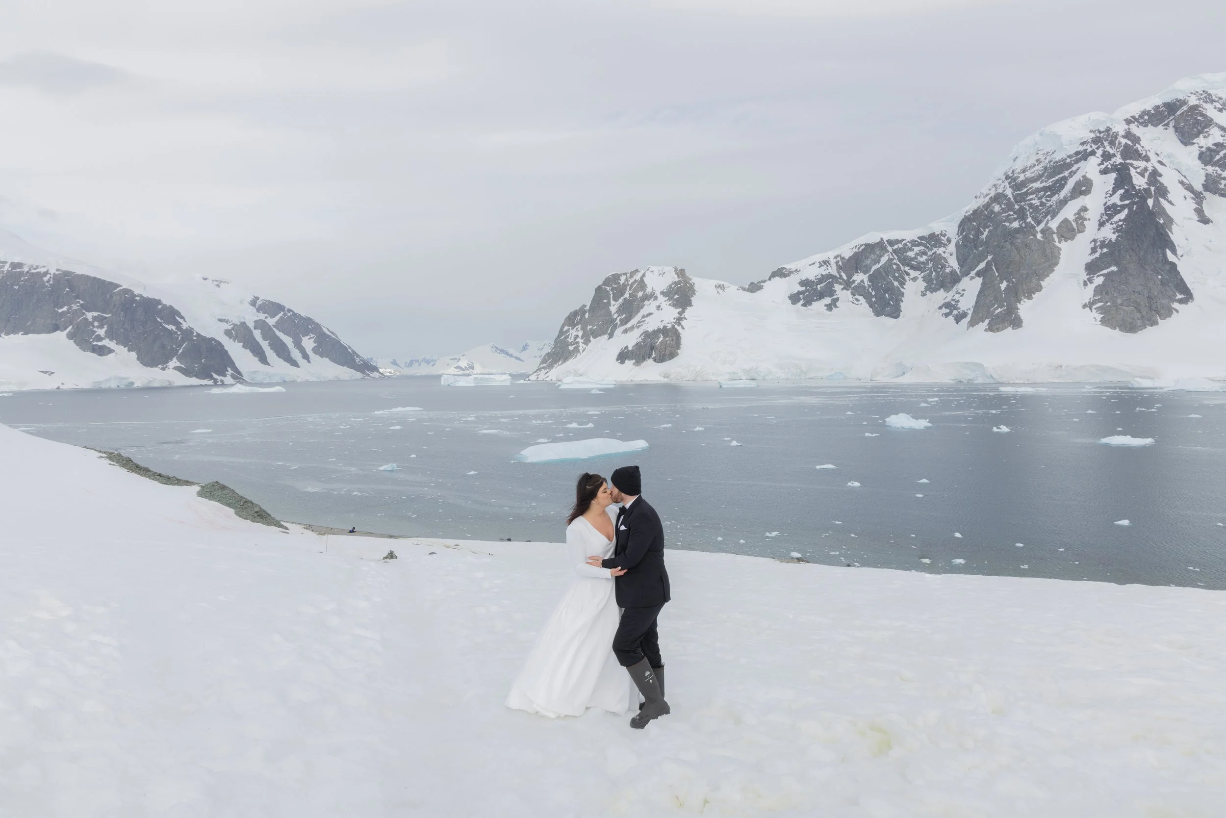 A bride and groom in wedding attire sharing a kiss in a snowy landscape with mountains, icebergs, and a body of water in Antarctica.