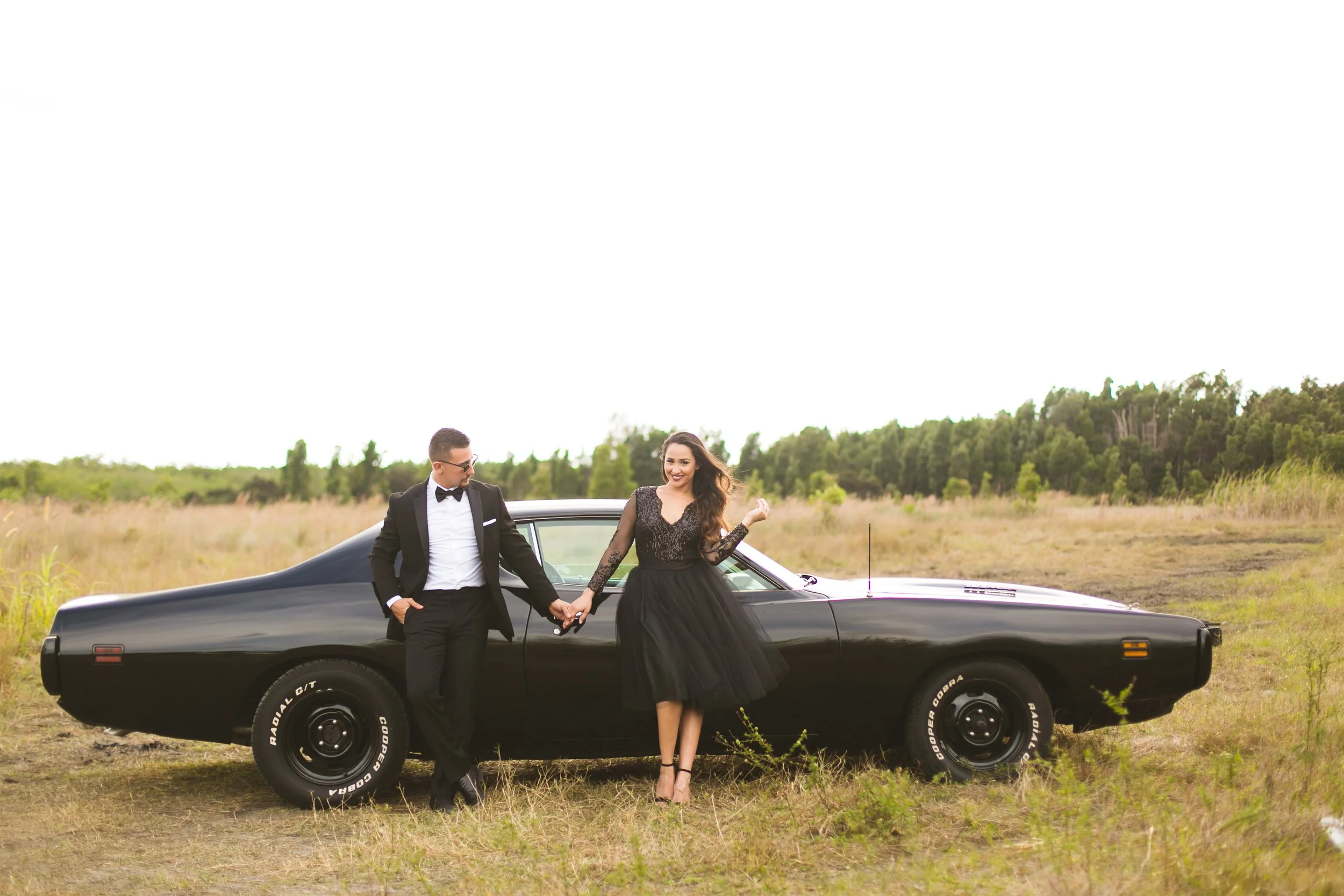 A man and woman in formal attire holding hands next to a classic black car in an open field with trees in the background.