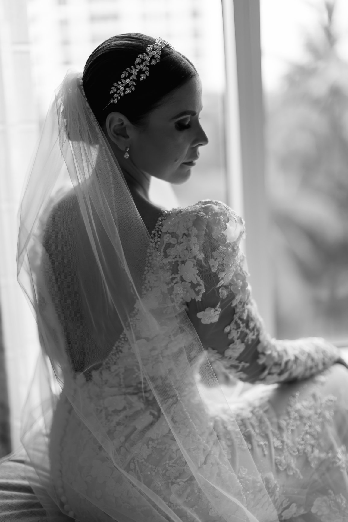 Black and white photo of a bride sitting by a window, wearing a lace wedding dress, veil, and a jeweled headband, looking down with a serene expression.