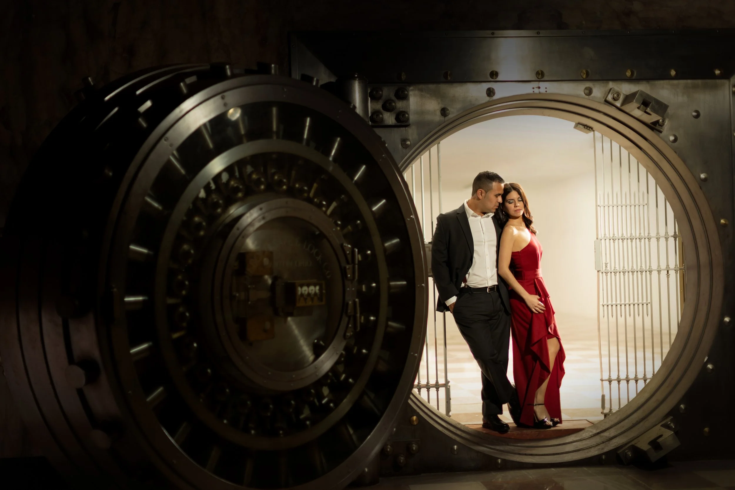 A man in a black suit and a woman in a red dress standing close together inside a large open bank vault, with their heads resting against each other.