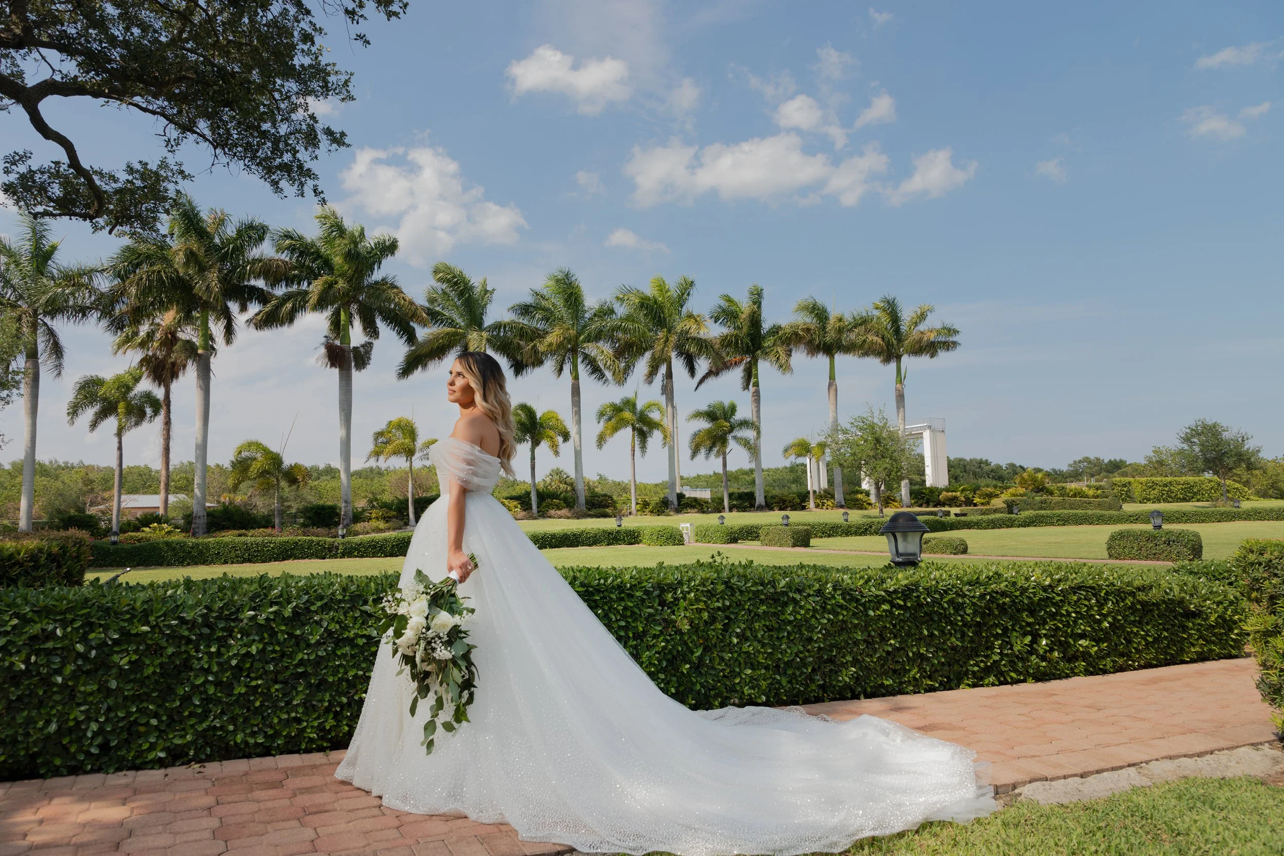 A woman in a flowy white wedding dress holding a bouquet of white flowers standing on a brick pathway in a lush green garden with palm trees and a blue sky.