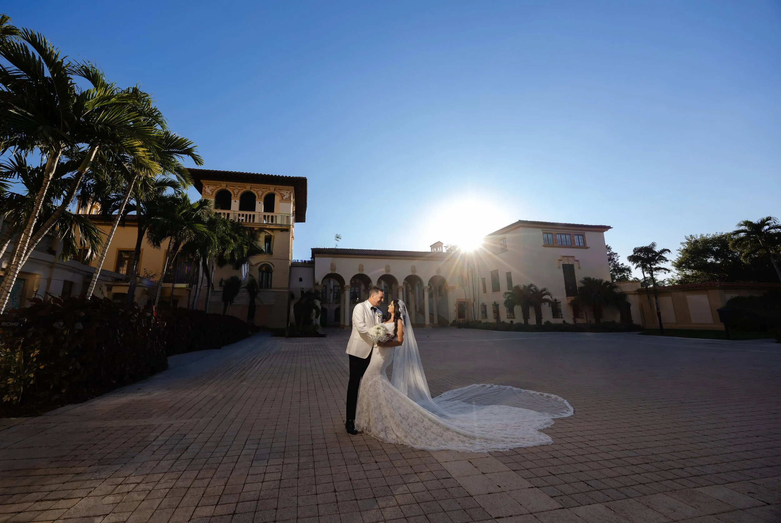 A bride and groom standing close together outdoors in front of a grand building at sunset, with the bride holding a bouquet and wearing a long lace wedding dress and veil.