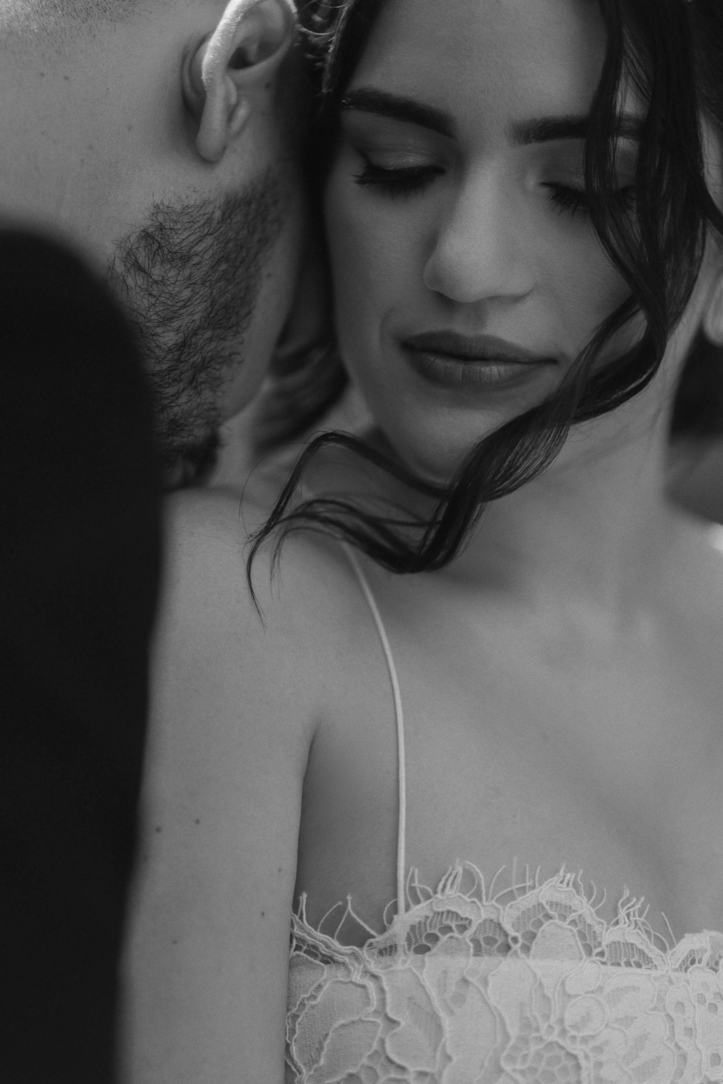 A black and white close-up photograph of a woman with closed eyes and dark hair with curls, wearing lace clothing, in an intimate pose with a man whose face is partially visible.