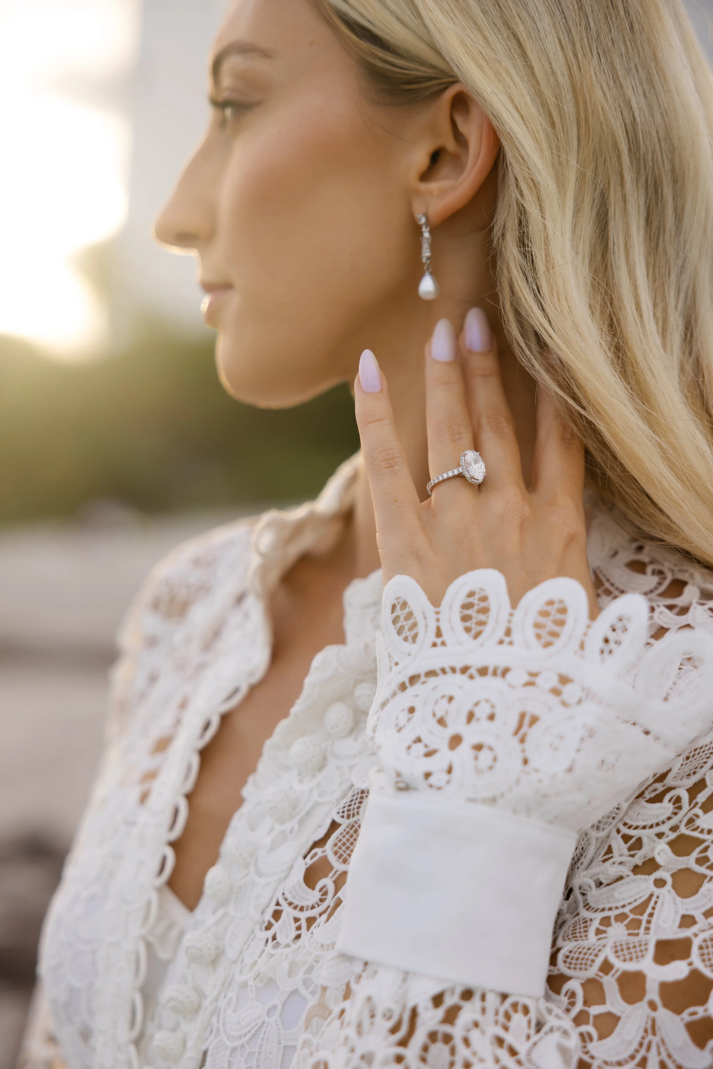 Close-up of a blonde woman with pearl drop earrings and a diamond ring on her hand, wearing a white lace dress, touching her earlobe and looking away outdoors.