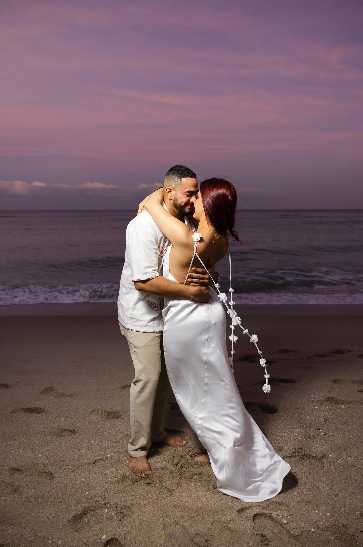 A couple embracing on the beach at sunset, the woman in a white dress and the man in a light shirt and pants, with purple and pink sky in the background.