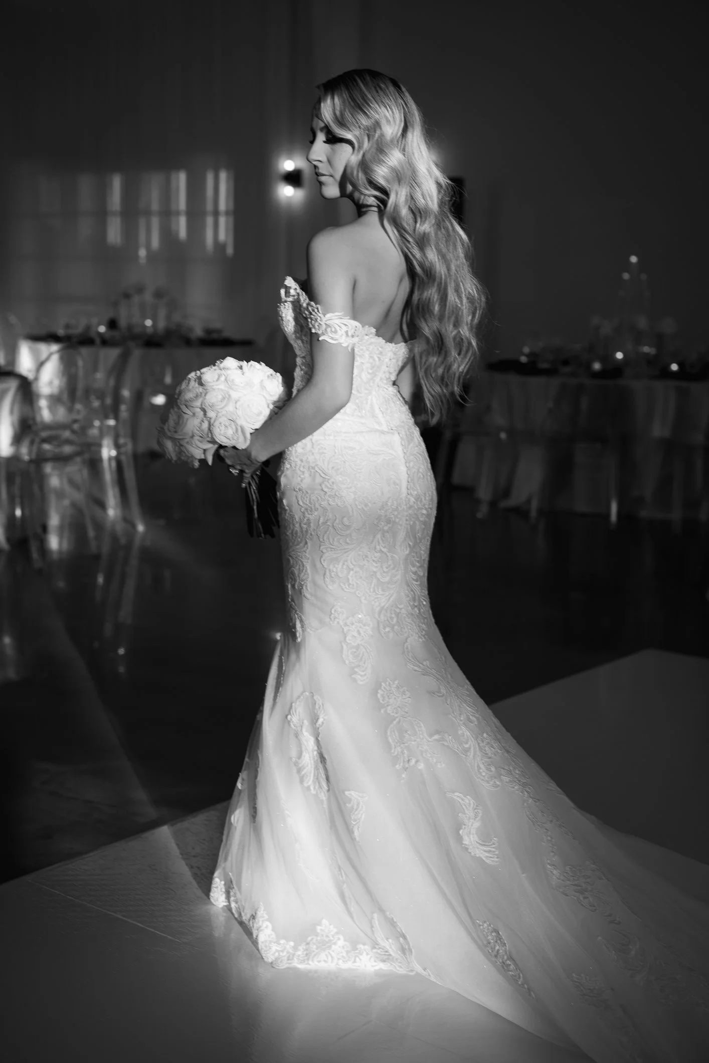 Black and white photo of a woman in a wedding dress holding a bouquet, inside a decorated venue with tables and chairs.
