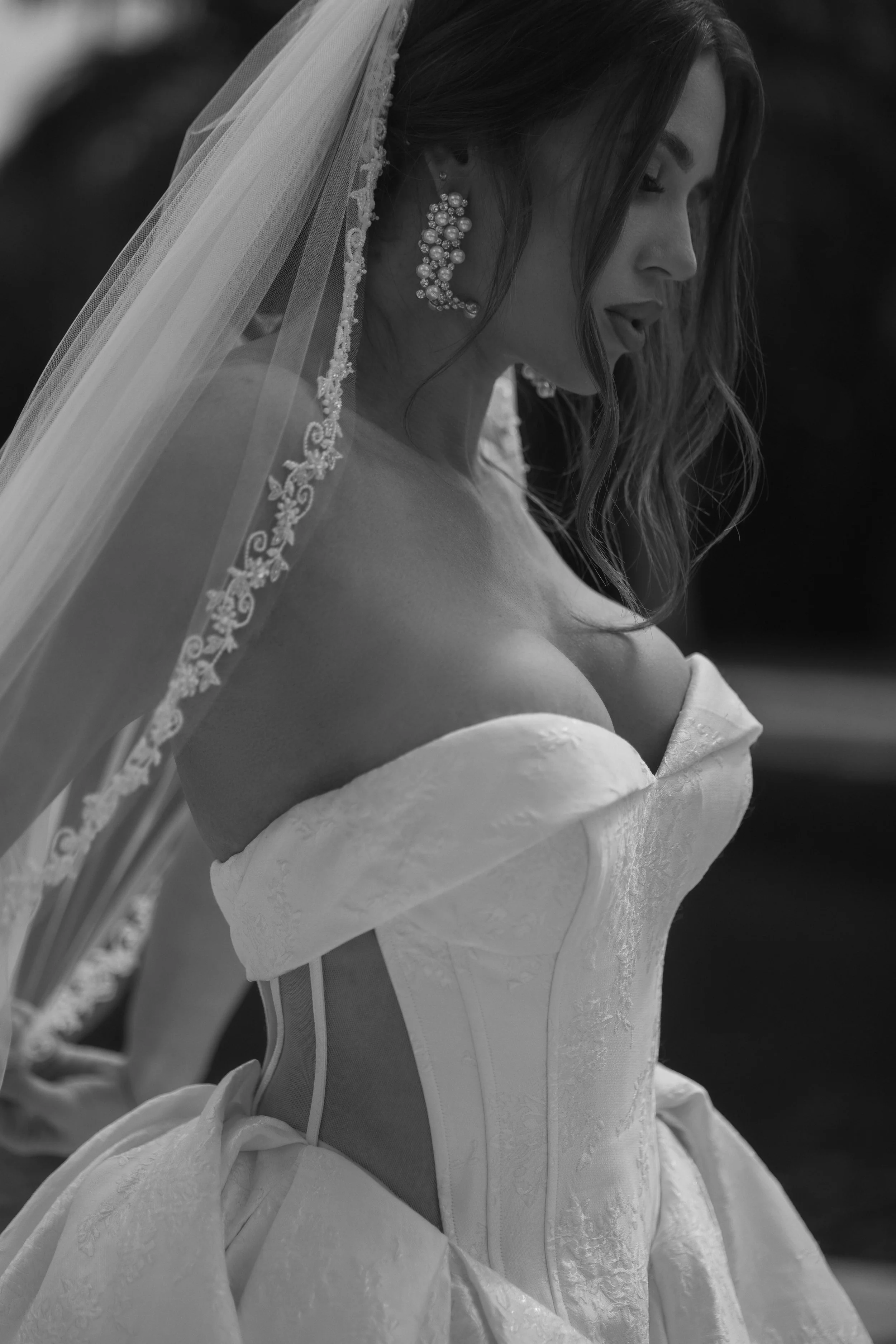 A woman in a wedding dress with off-the-shoulder sleeves, wearing a veil and large pearl earrings, looking down with her eyes closed.