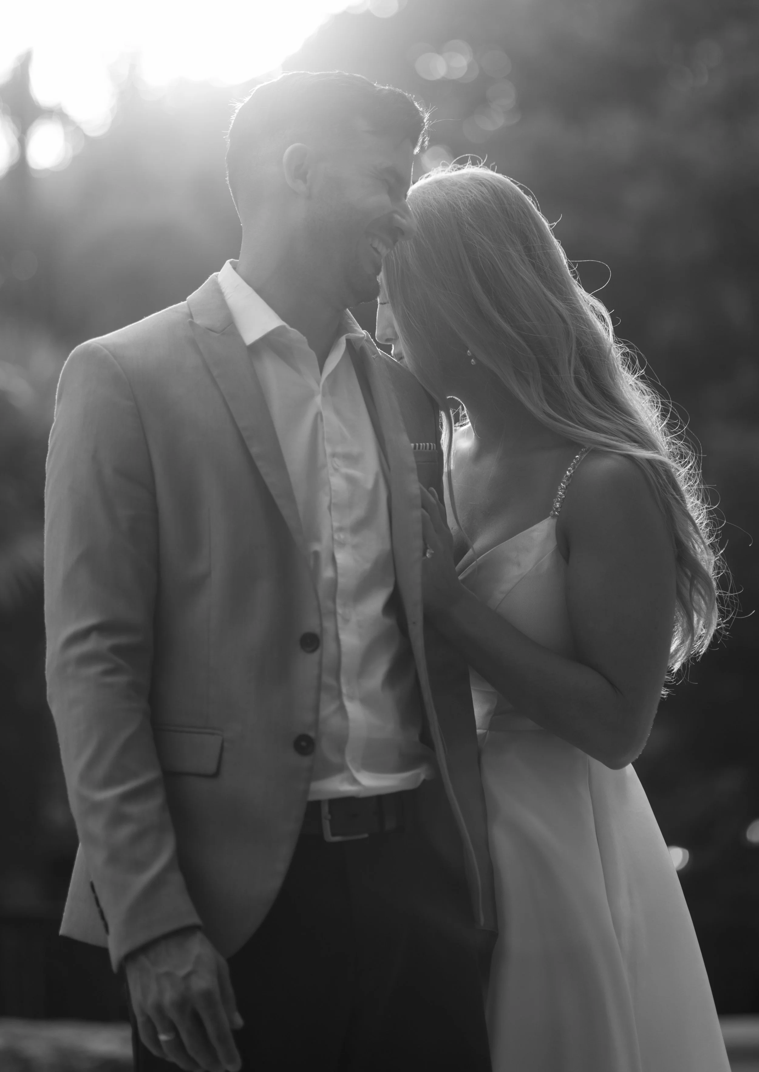 A black-and-white photo of a couple in formal attire, smiling and embracing outdoors with sunlight behind them.
