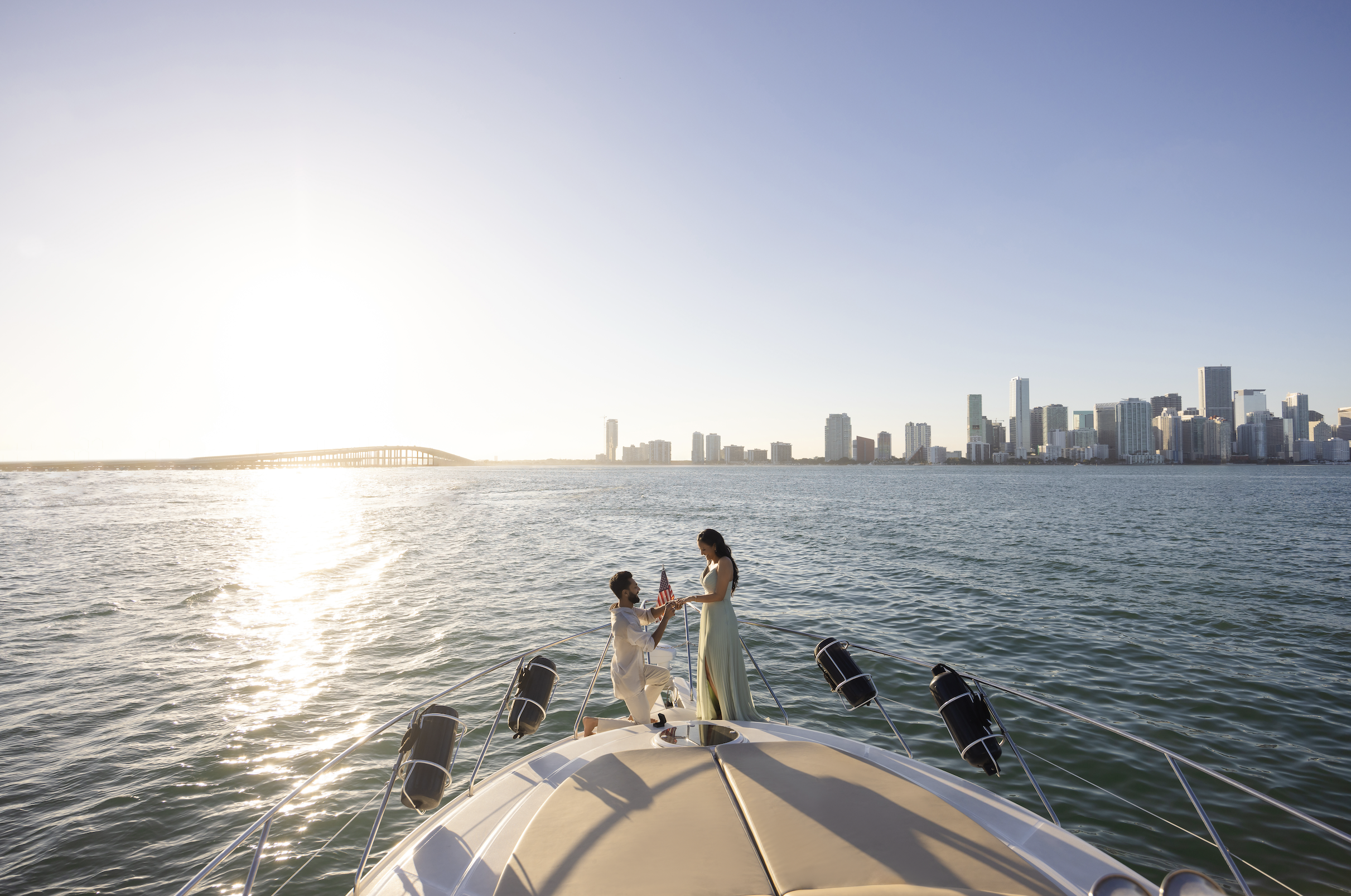 A couple on a yacht at sunset, with a city skyline in the background. The man is proposing to the woman.