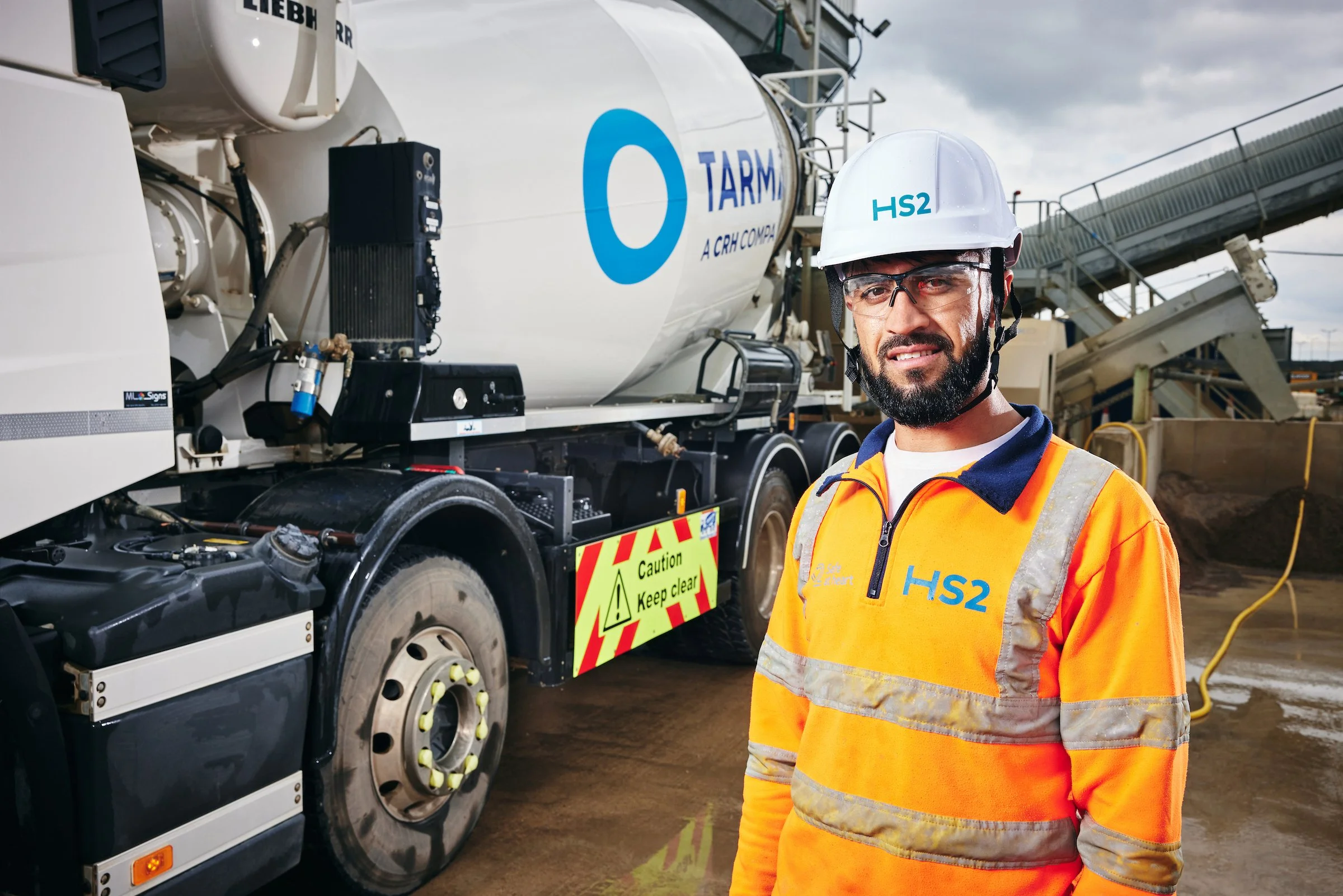 Construction worker wearing a white helmet and orange safety vest standing in front of a large cement truck at a construction site.