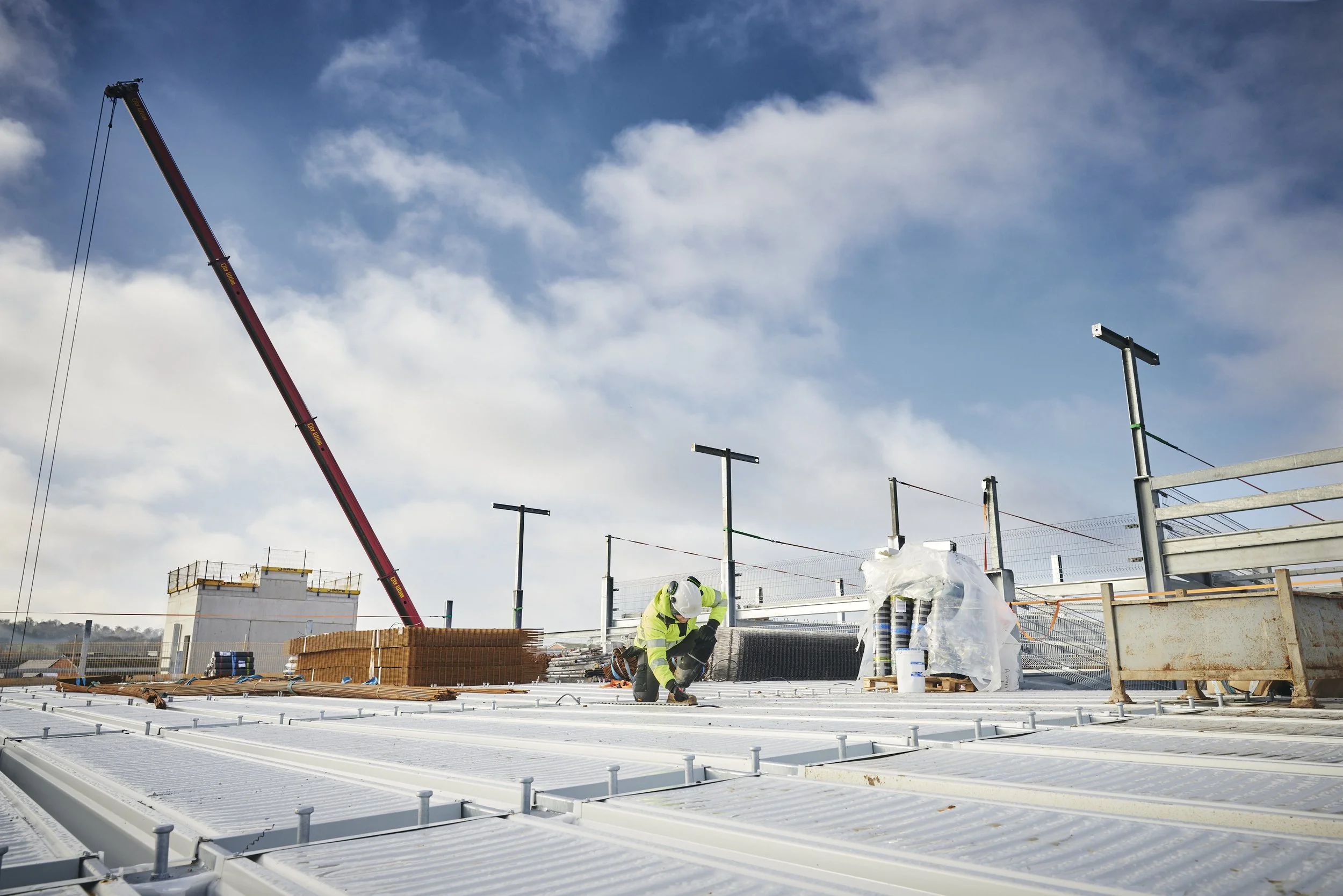 Construction worker in safety gear kneeling on roof during building construction, with construction materials and a crane in the background under a blue sky with clouds.