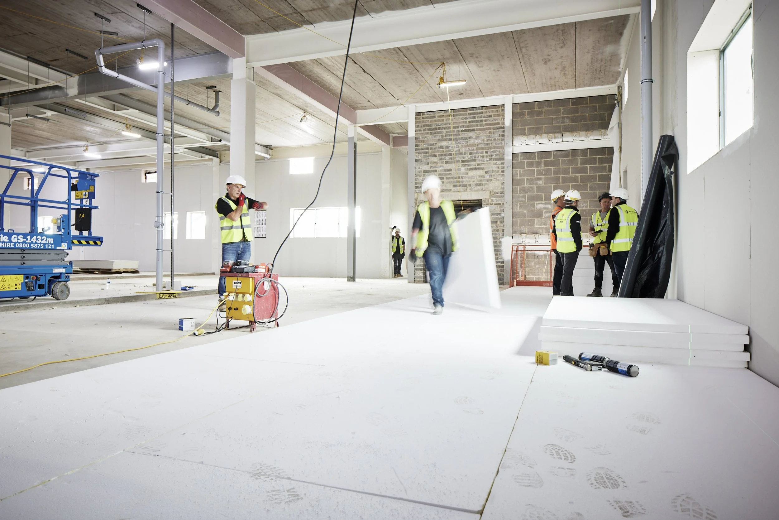 Construction workers in a large indoor space with exposed ceilings and brick walls, some wearing safety vests and helmets, working on flooring and materials.