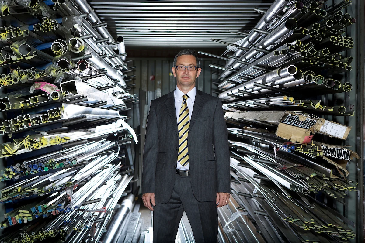 A man in a business suit standing in a storage area with metal pipes and tubes stored on shelves on both sides.
