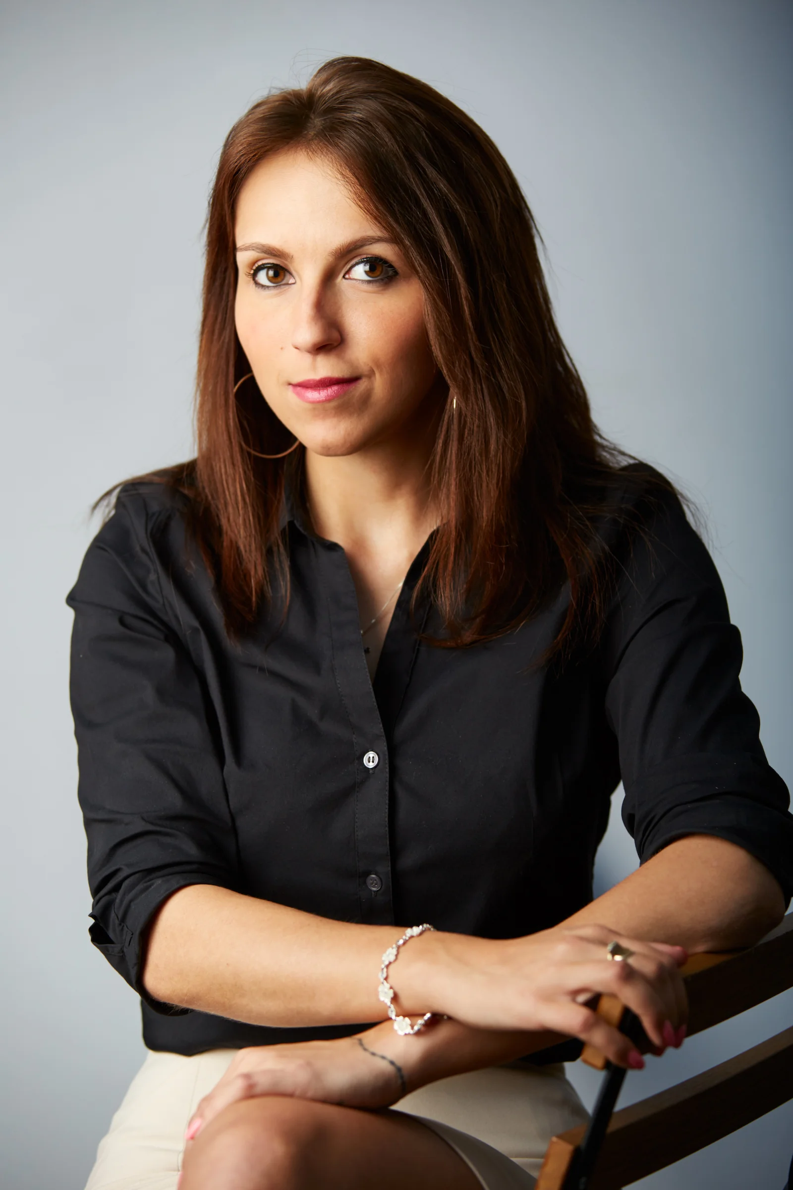 A woman with brown hair and light skin wearing a black blouse and jewelry, sitting with her arms crossed on a wooden chair, against a plain grey background.