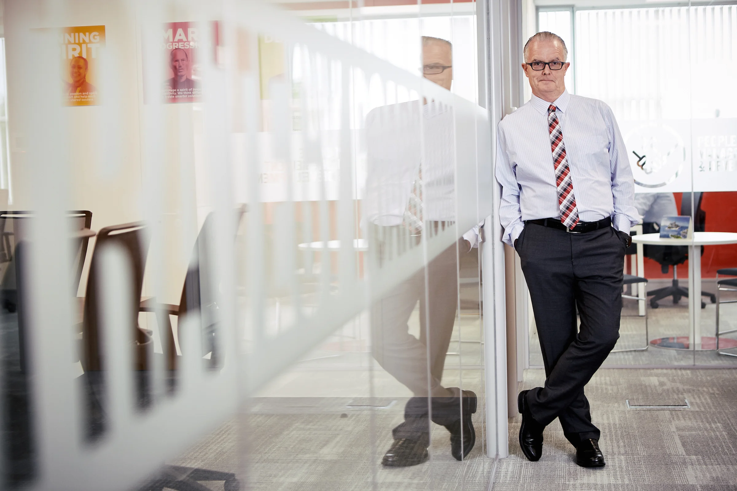 A middle-aged man in a white dress shirt, striped tie, and dark pants leaning against a glass wall in an office. His hands are in his pockets, and he is wearing glasses with his hair slicked back. Behind him, there is a meeting room with chairs, post