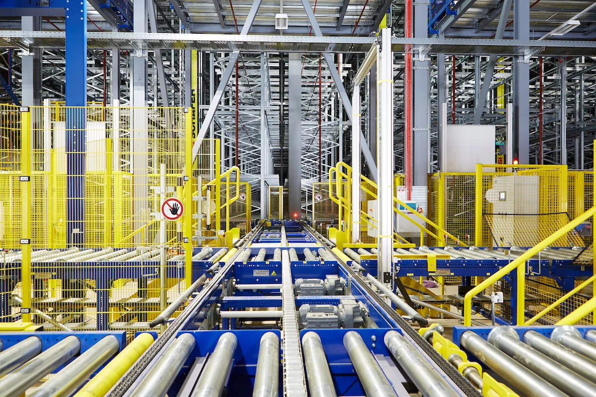 Industrial warehouse with blue and yellow machinery and conveyor tracks, safety fencing, and metal shelving extending into the distance.
